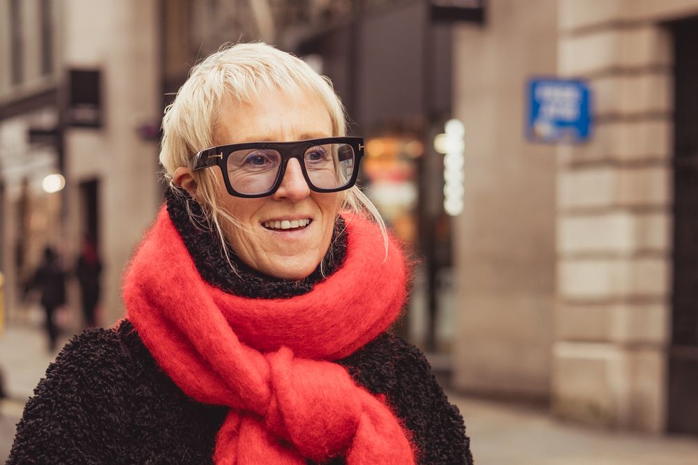 Person wearing black-rimmed glasses and a bright red scarf smiles warmly on a city street.