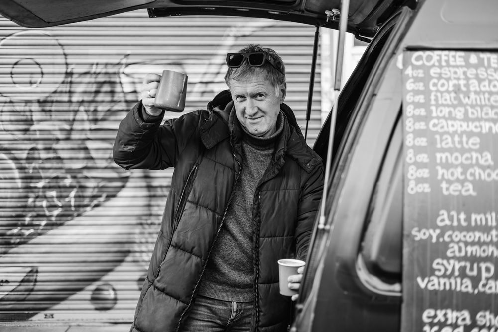 A person in a puffy winter jacket gestures while standing next to a coffee truck with a menu board visible in black and white.