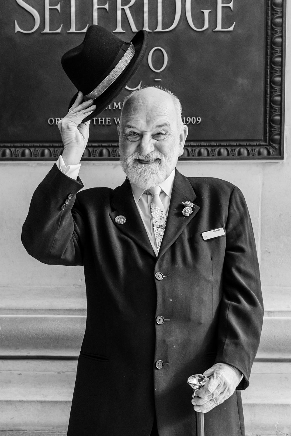 Black and white vintage photograph of a bearded gentleman in a suit tipping his hat in front of a Selfridges sign.