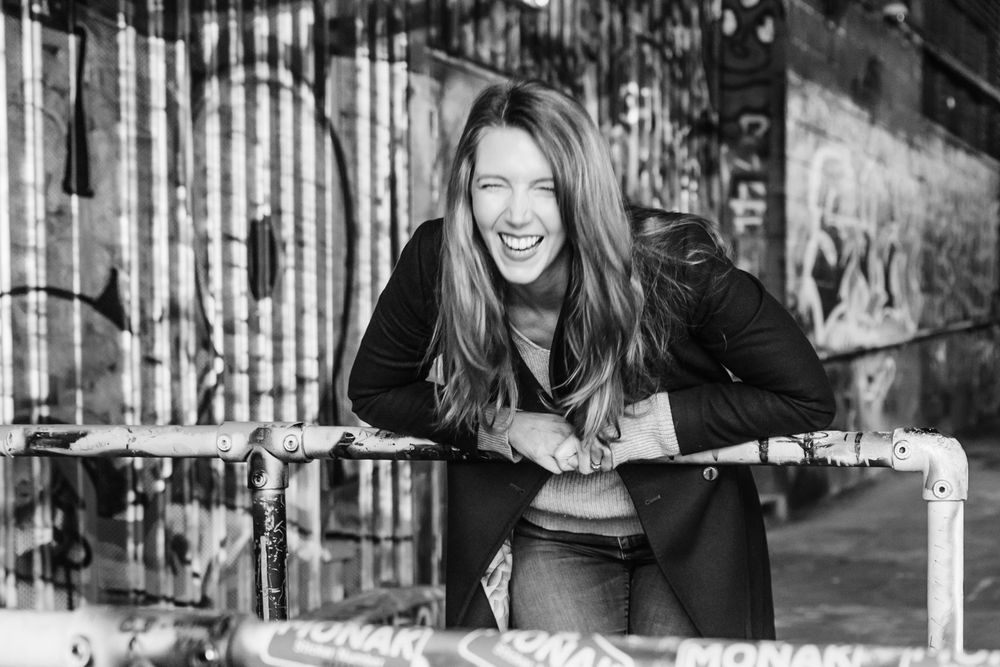 A joyful person in black clothing leans on a metal railing while laughing outdoors in a black and white photo.