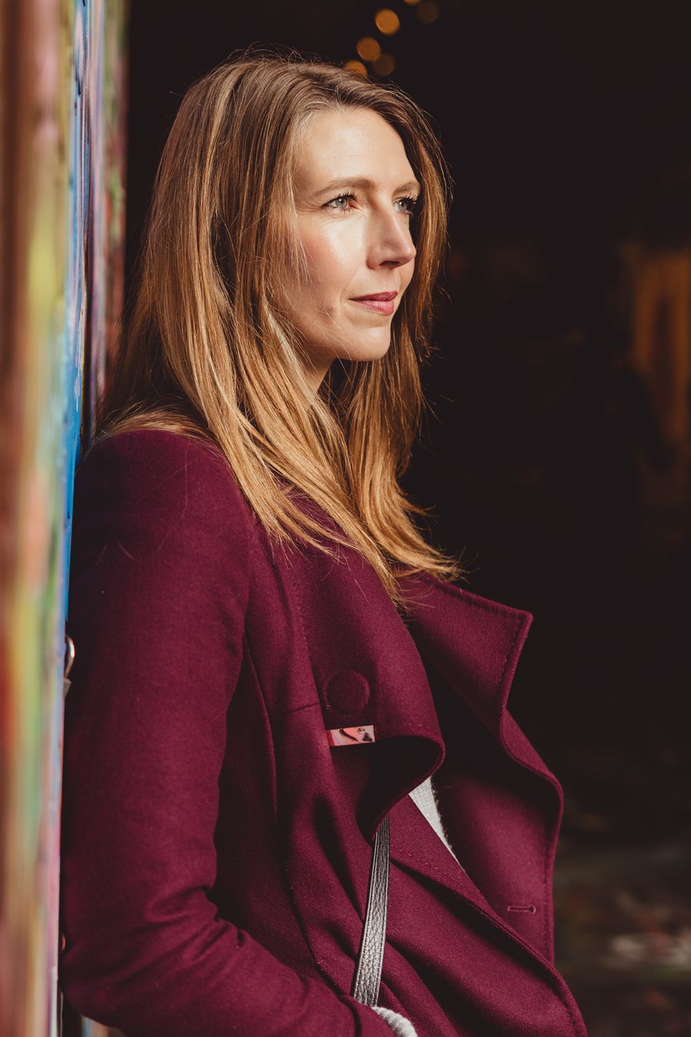 Portrait of someone wearing a burgundy coat looking to the side with soft lighting and long blonde hair.