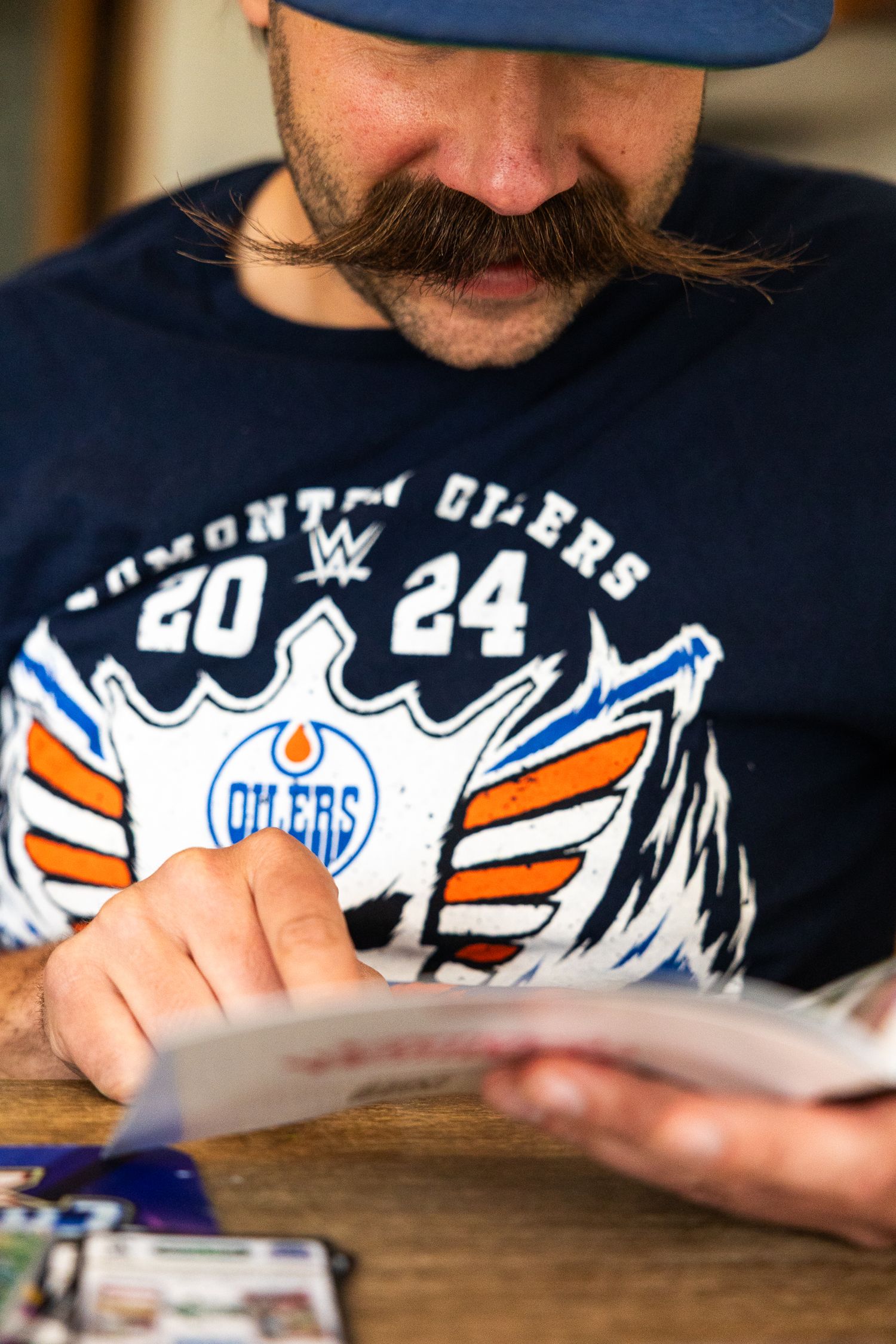 A man with a handlebar mustache wearing an Edmonton Oilers shirt reads trading cards.