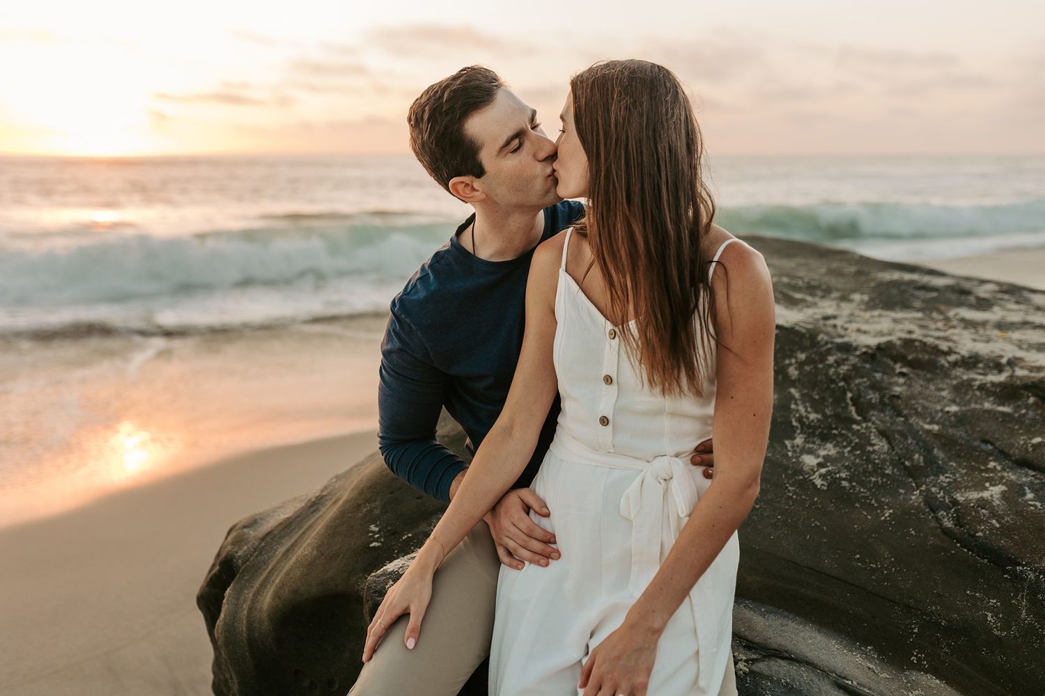A romantic couple shares an intimate moment at Windansea Beach at sunset, with waves crashing against rocky shore in background.