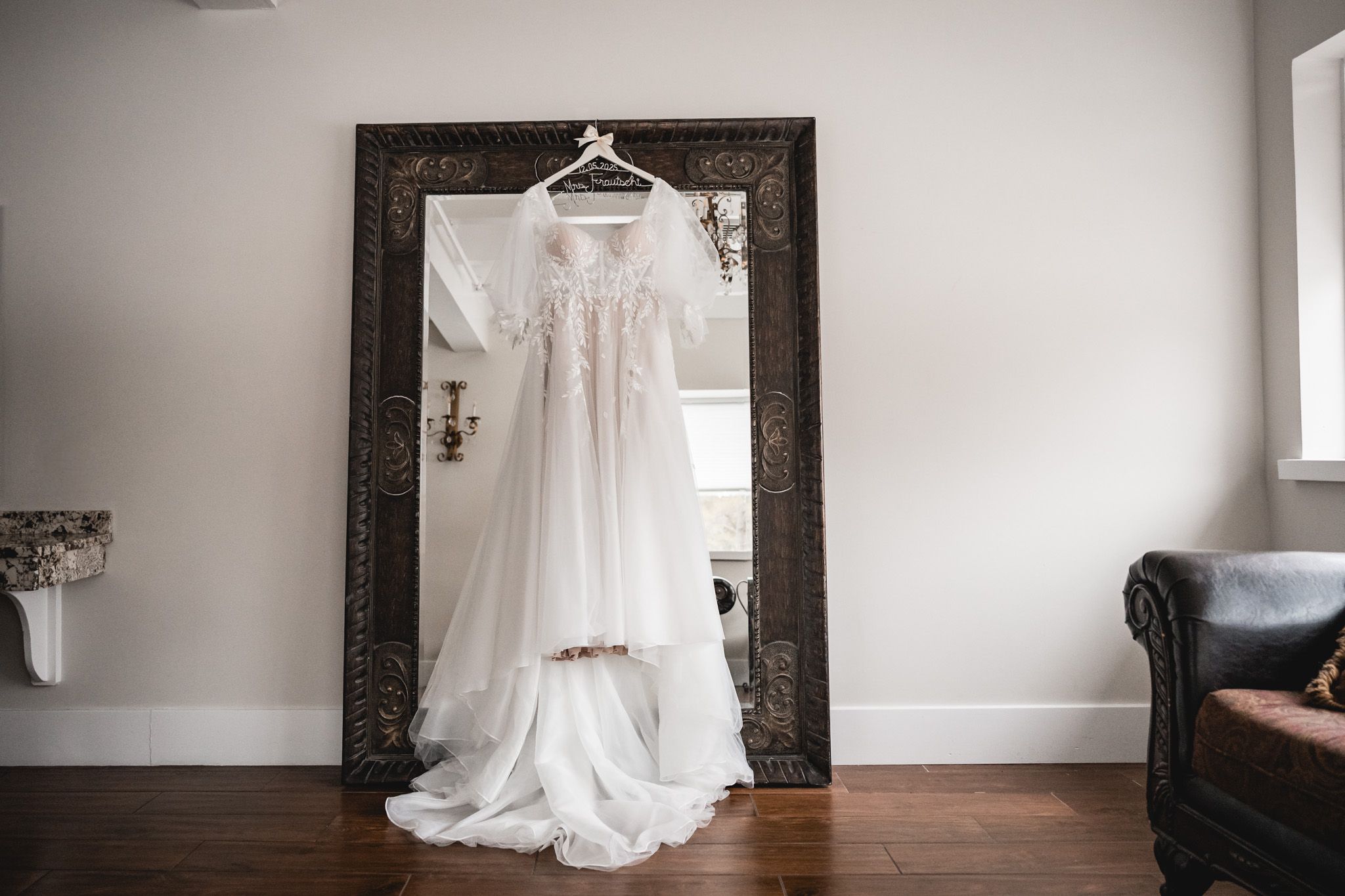 White wedding dress hanging on display against an ornate dark frame mirror.