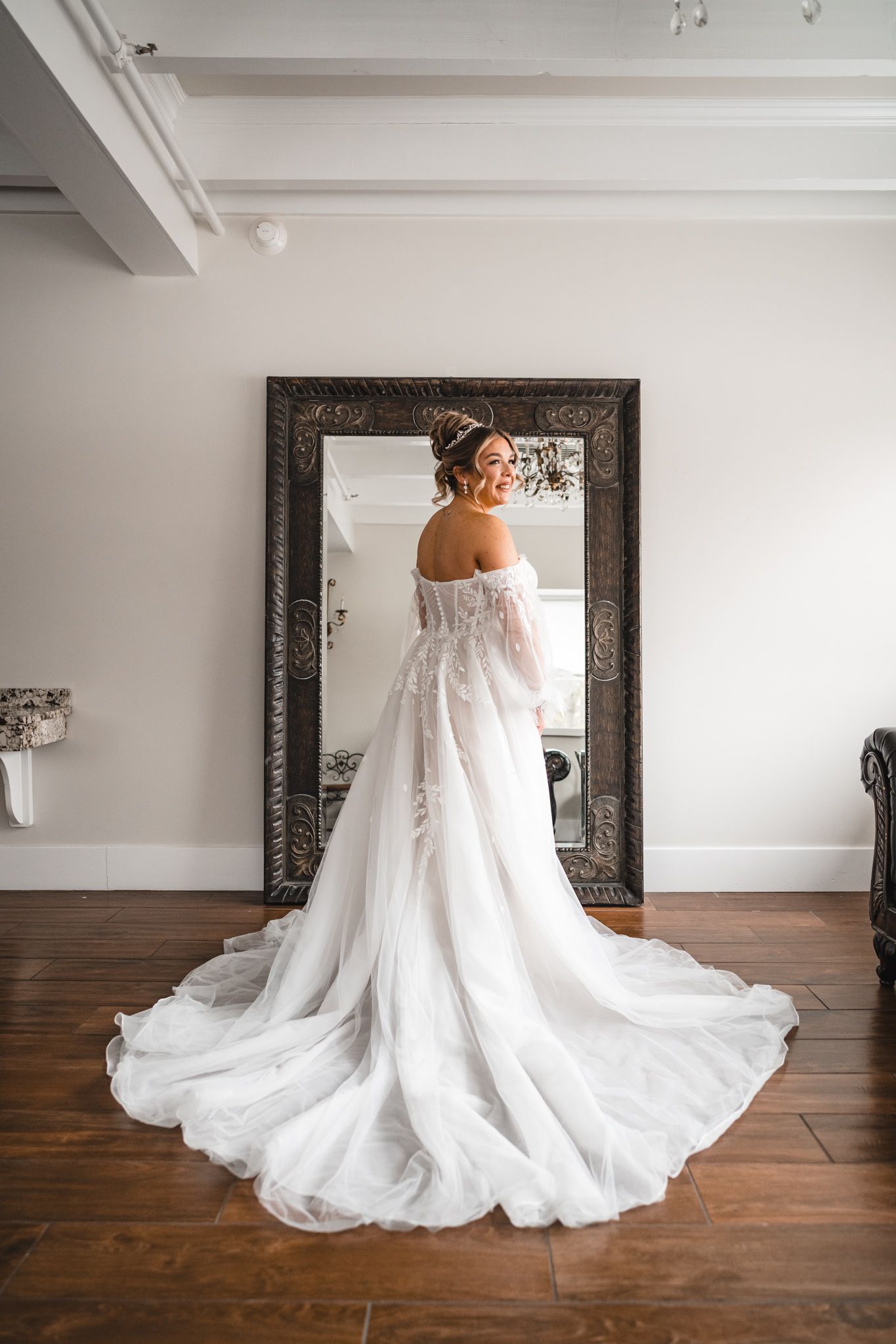 Elegant wedding gown with flowing train displayed on hardwood floor with ornate mirror.