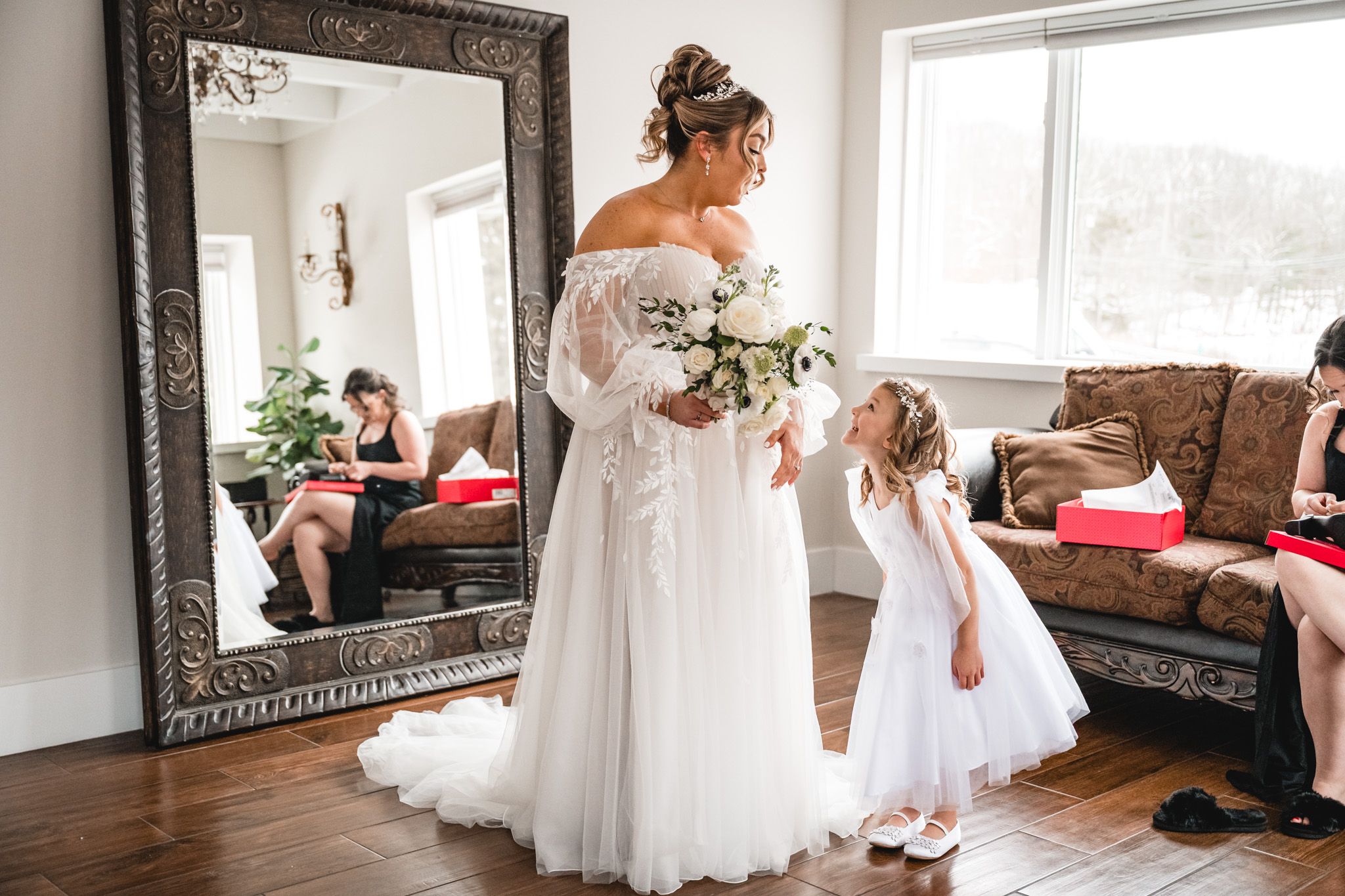 A bride in an off-shoulder lace gown stands with a flower girl in white near an ornate mirror in a living room.
