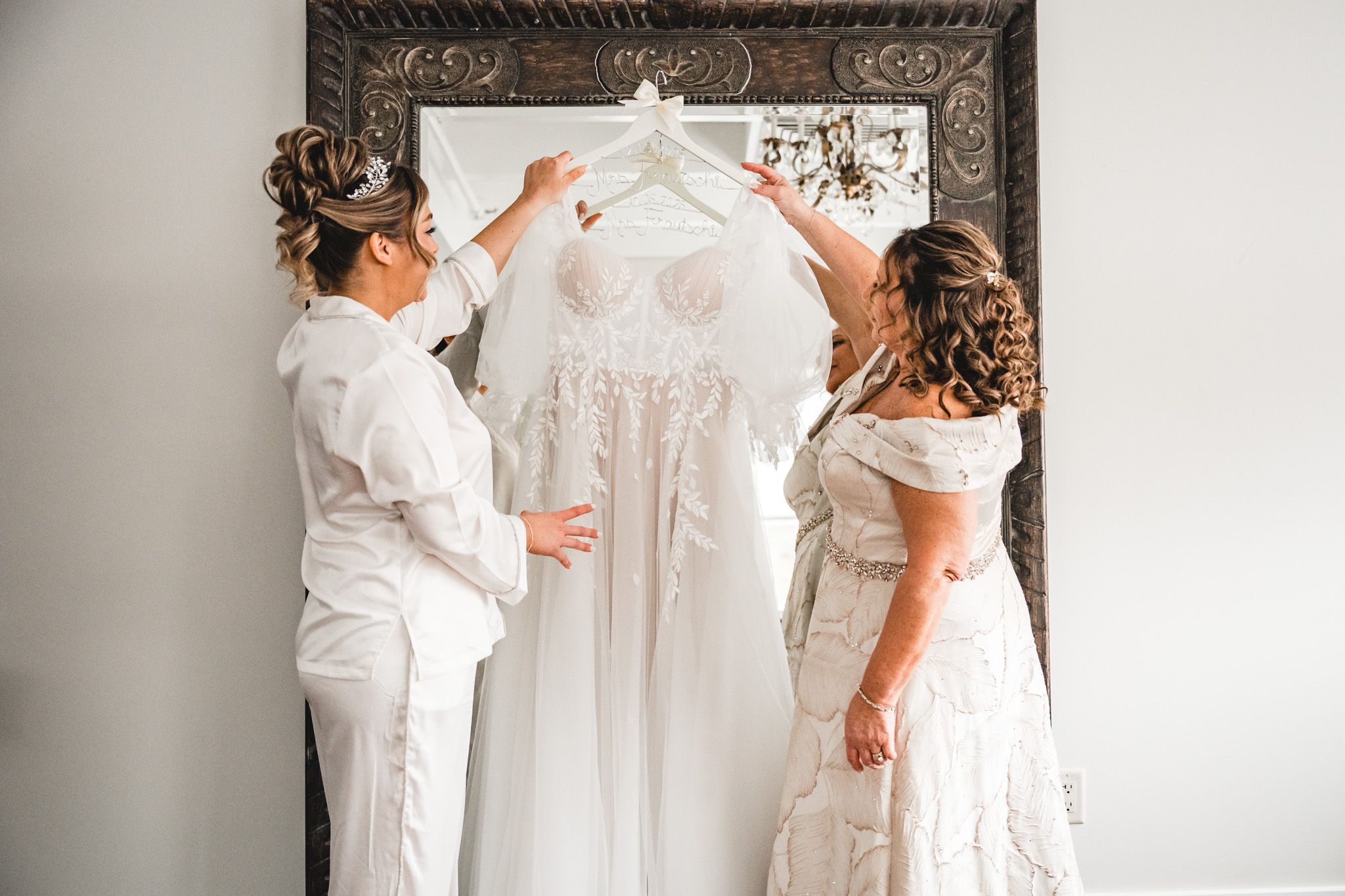 Friends helping with wedding dress preparations in a bright room with ornate mirror.