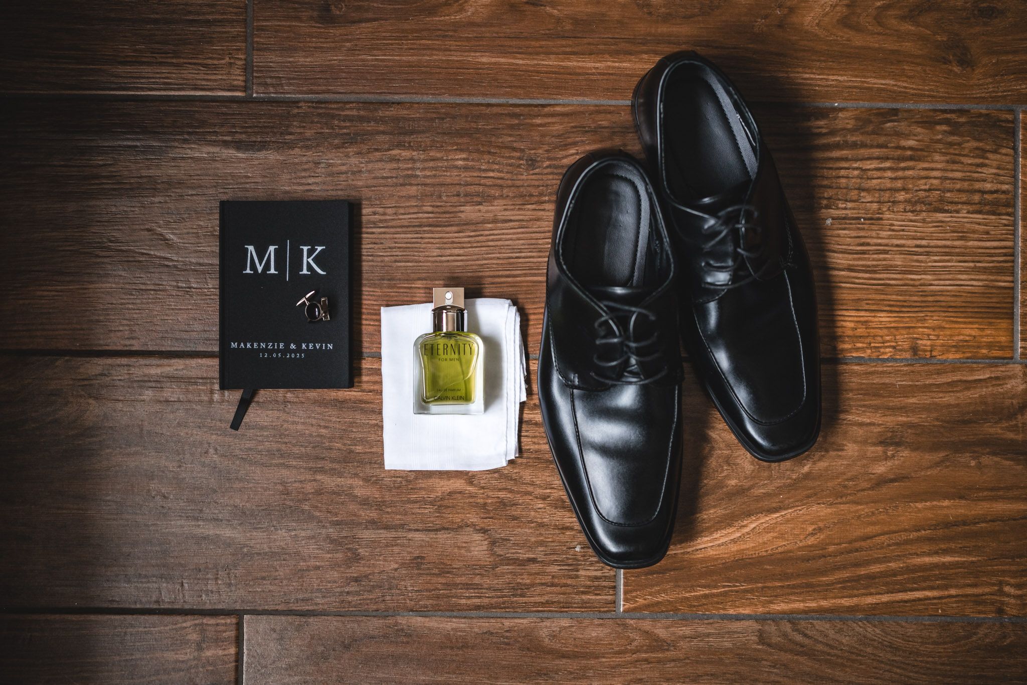 Groom's accessories including black dress shoes, cologne and a monogrammed black book on wooden floor.