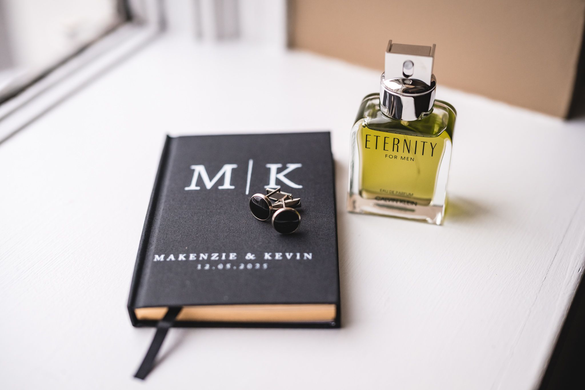 A black monogrammed book and bottle of cologne arranged on a surface near a window.