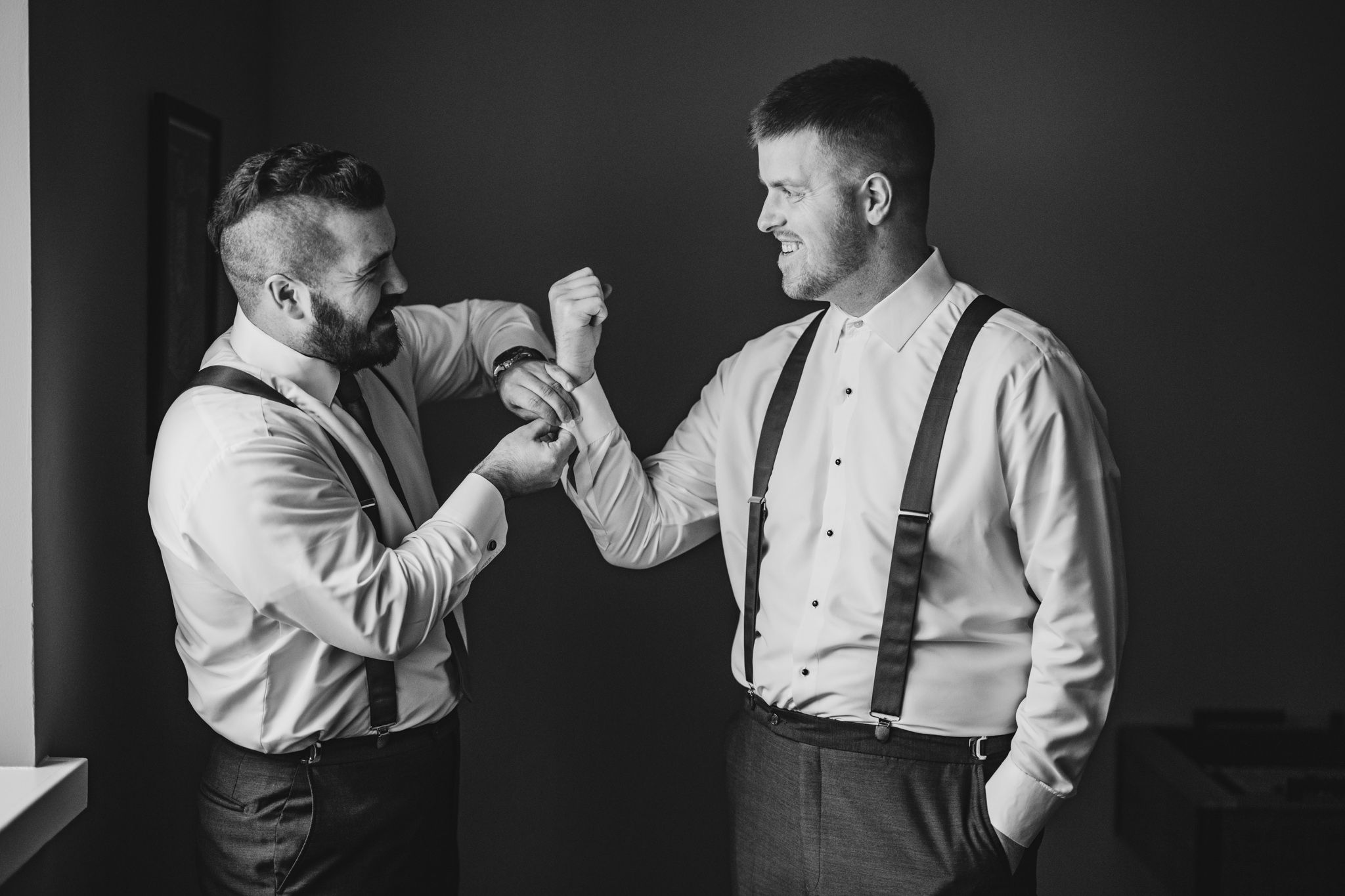 Two men in formal attire and suspenders share a lighthearted moment while getting ready.