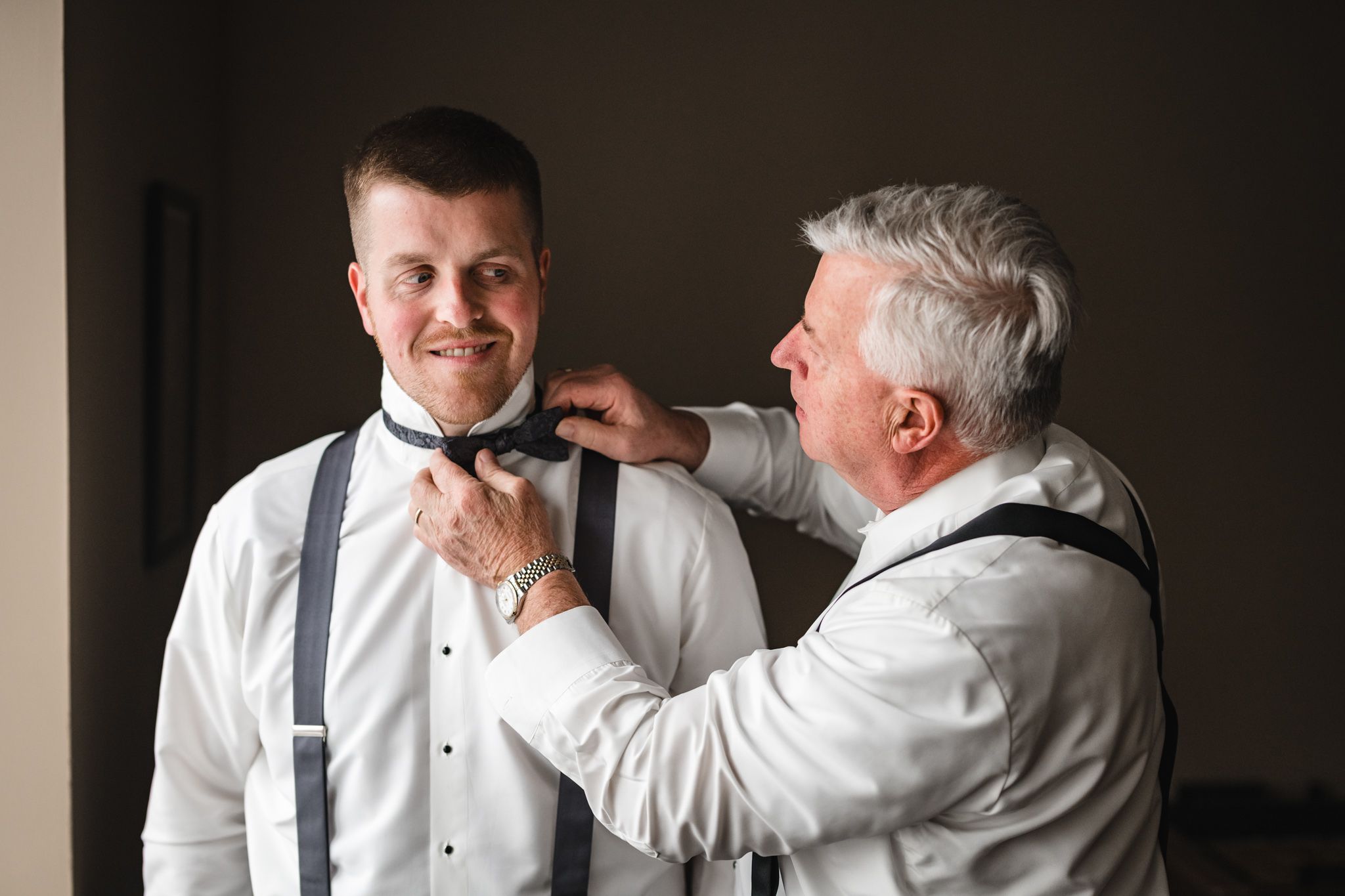 An older man helps adjust the bow tie of a younger man in formal attire.