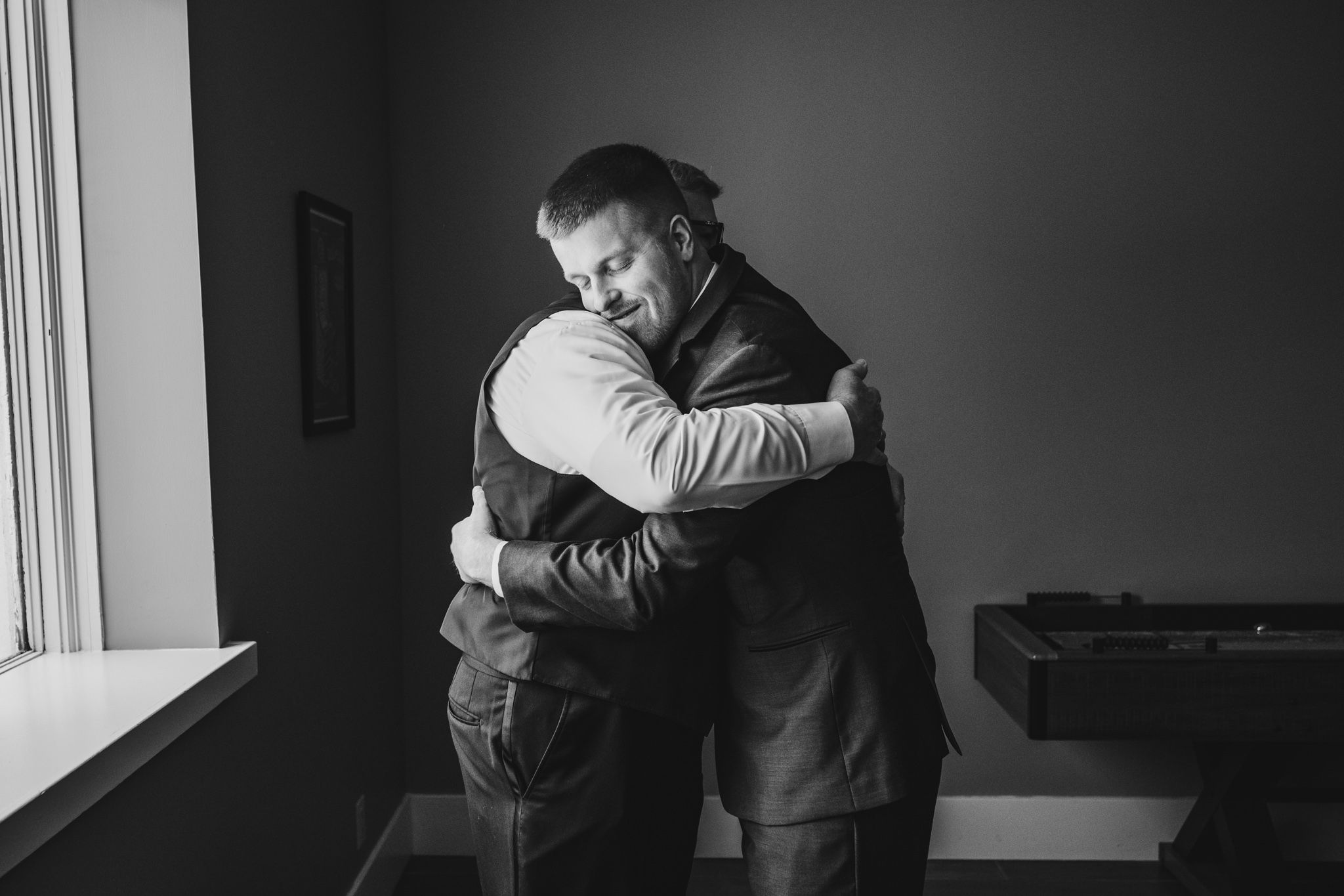 Two men share an emotional embrace near a window in formal wedding attire.