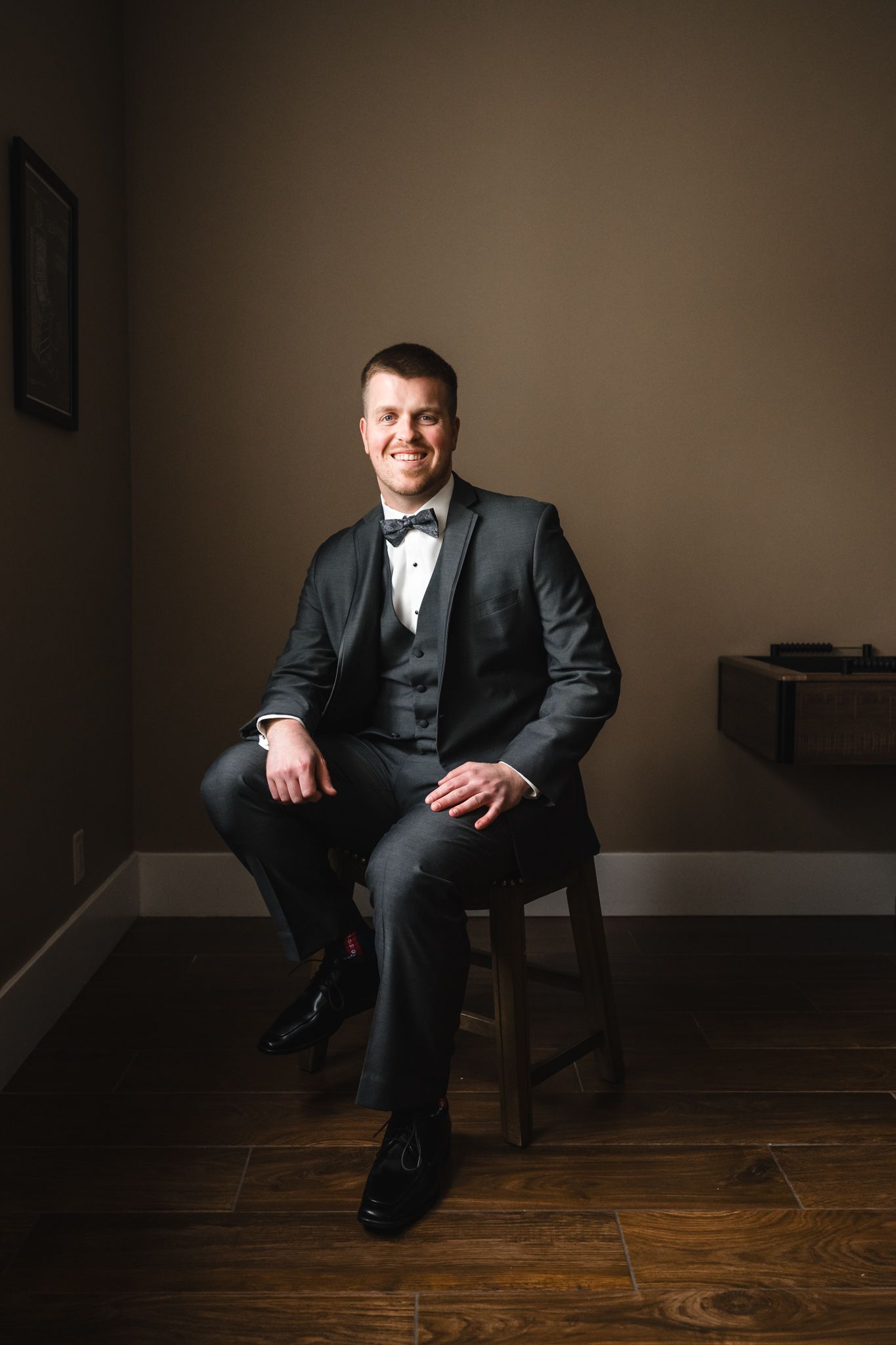 Groom in black tuxedo and bow tie poses in dramatic lighting against dark background.