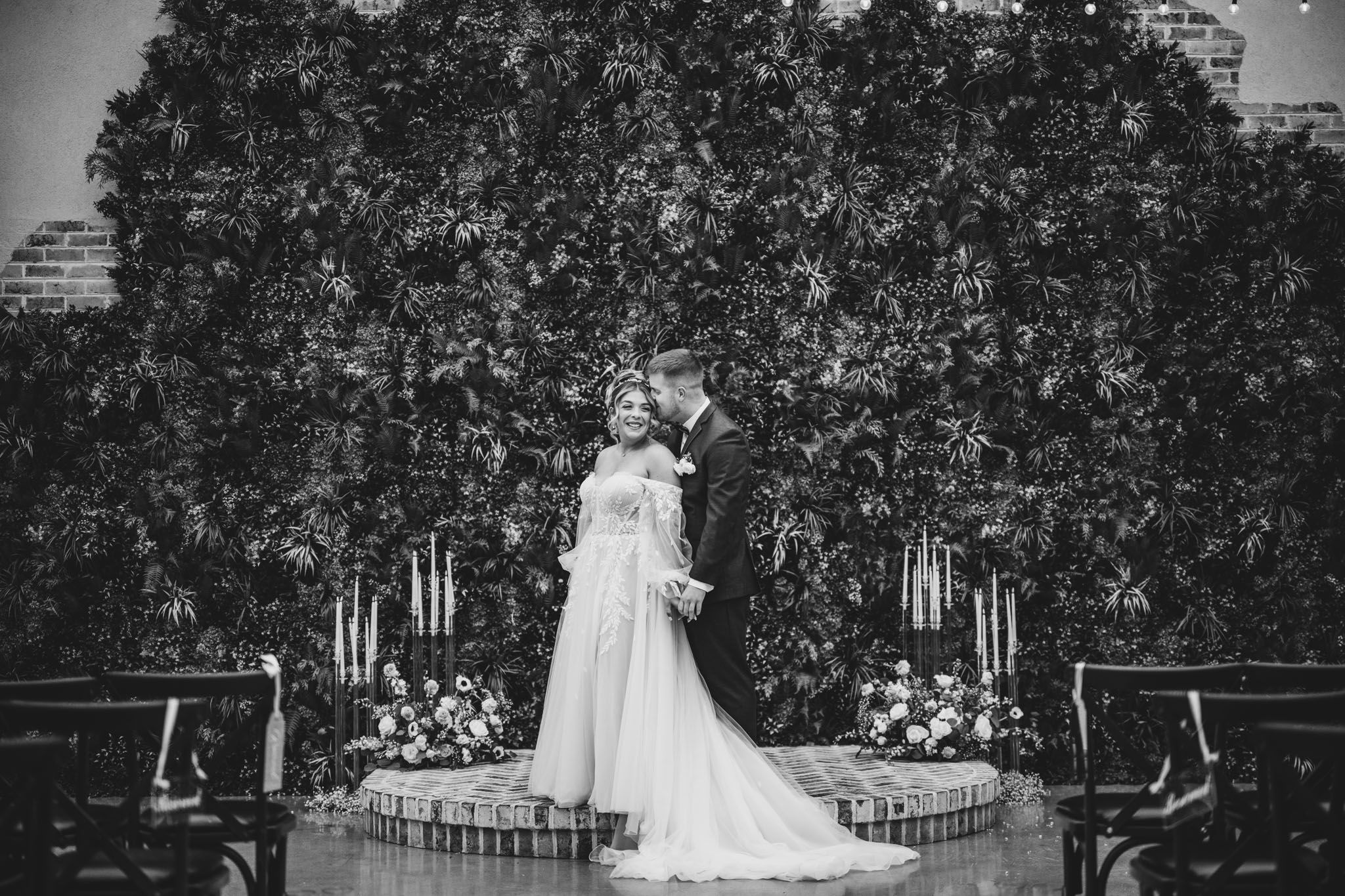A black and white wedding photo with a couple standing before an elegant floral wall backdrop.