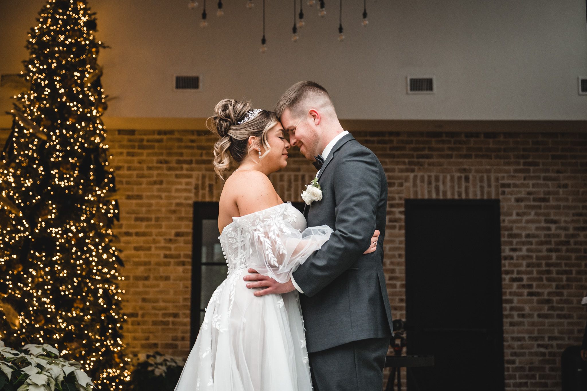 A festive wedding moment captured beside a glowing Christmas tree with exposed brick walls.