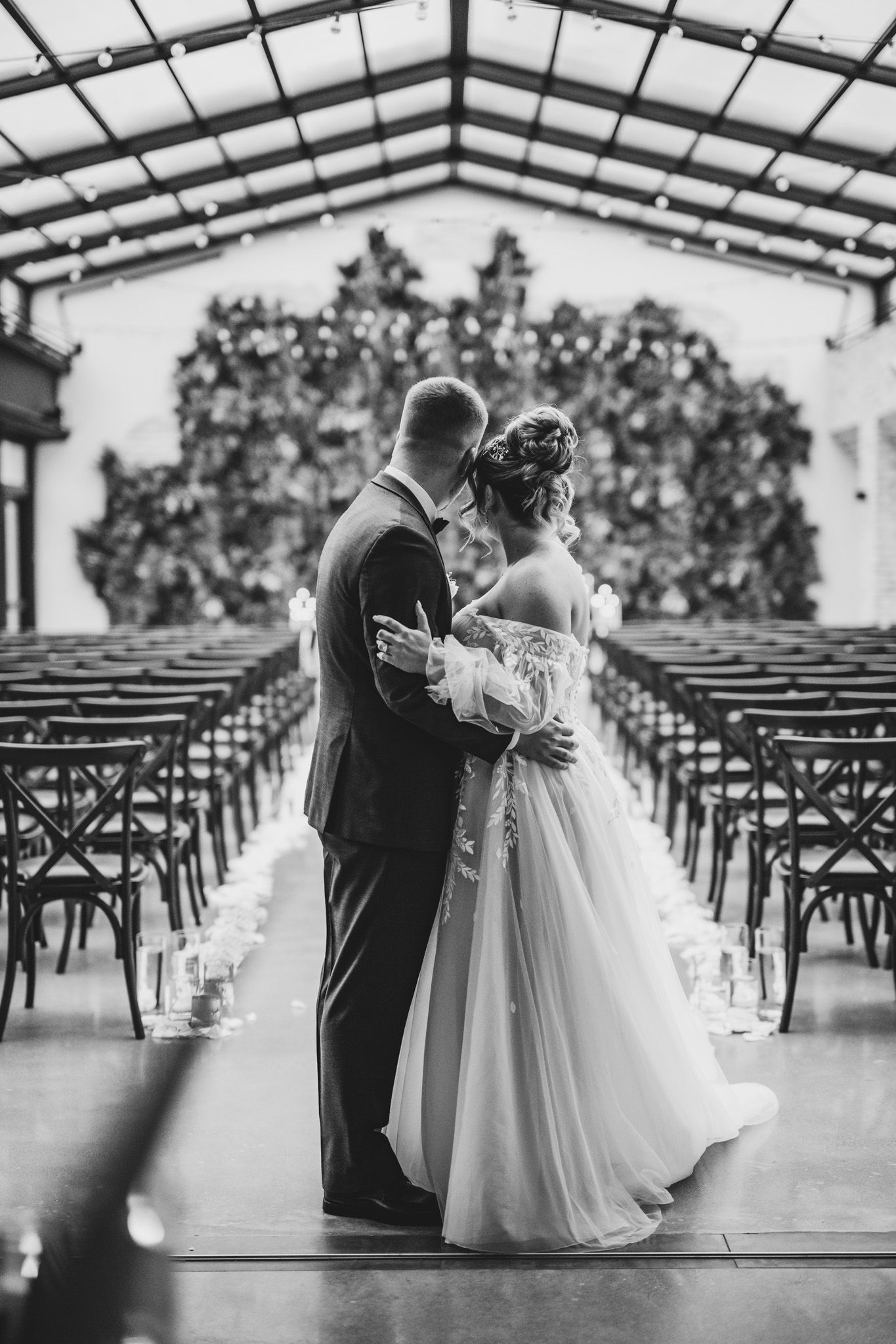 A black and white wedding photo in an elegant venue with rows of chairs and a floral arch backdrop.