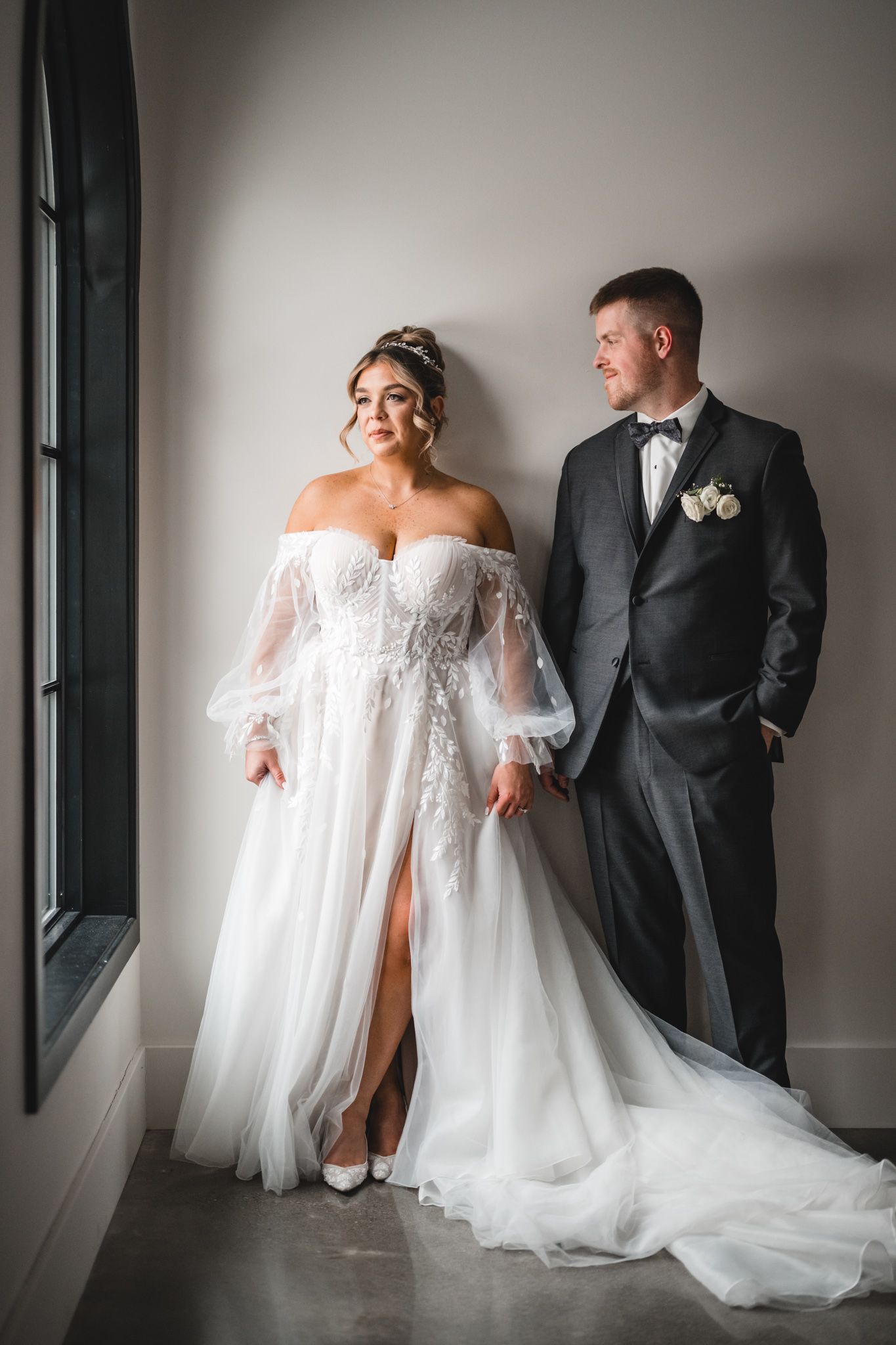 An elegant wedding portrait by a window featuring a flowing off-shoulder gown with sheer sleeves.