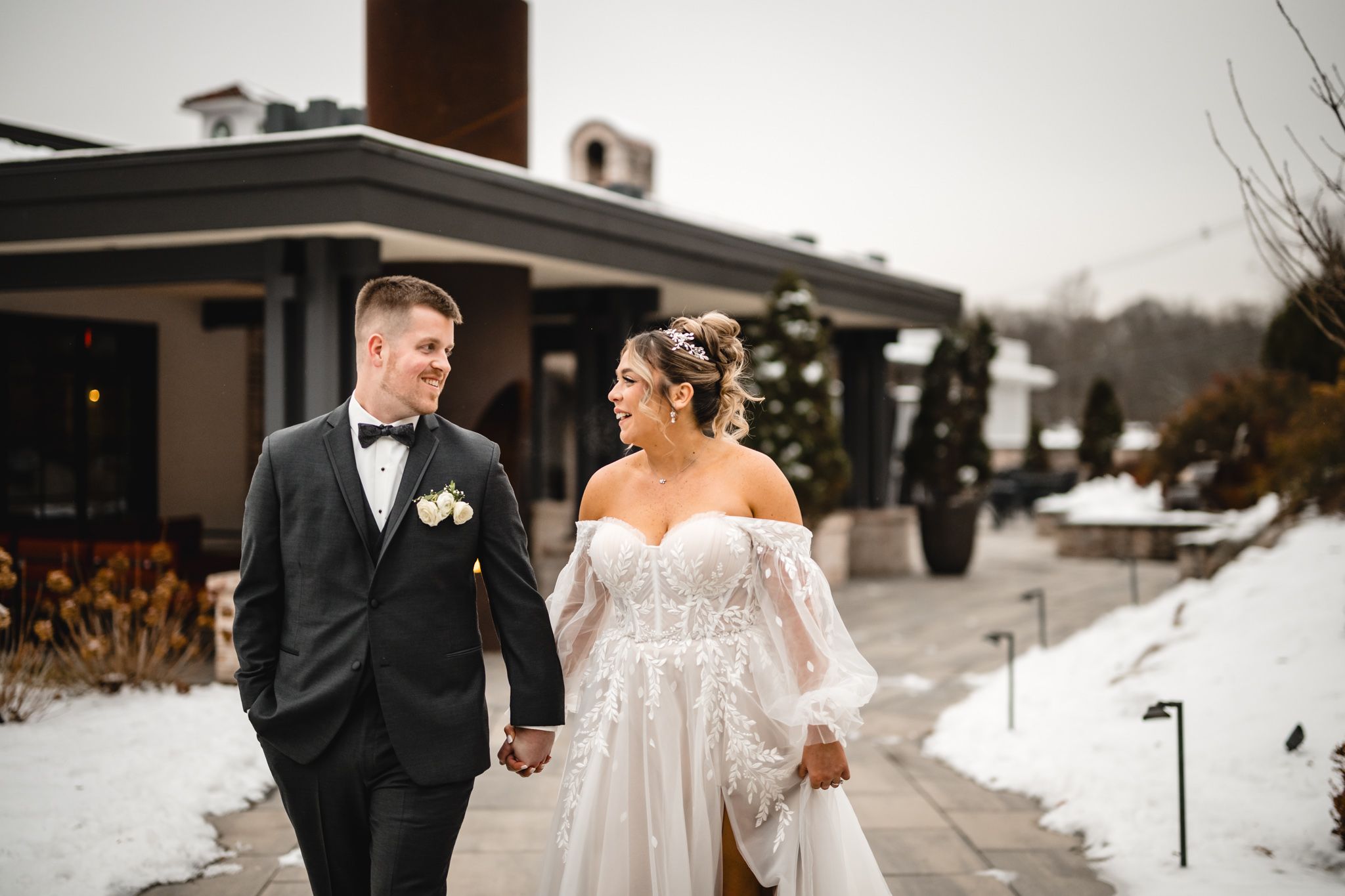A winter wedding photo with snow-covered grounds and modern architecture in the background.