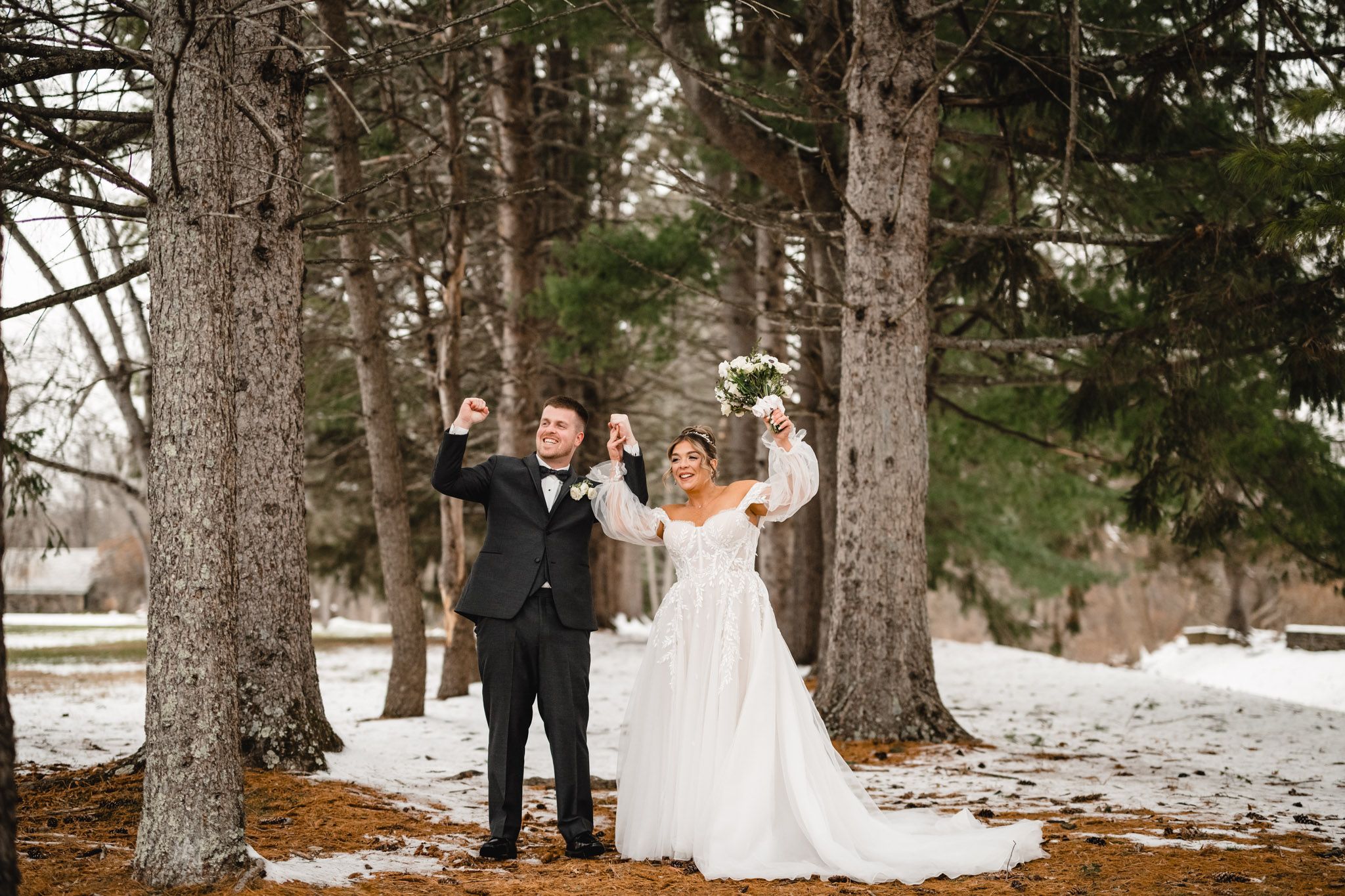 Wedding couple poses between tall pine trees in winter setting.