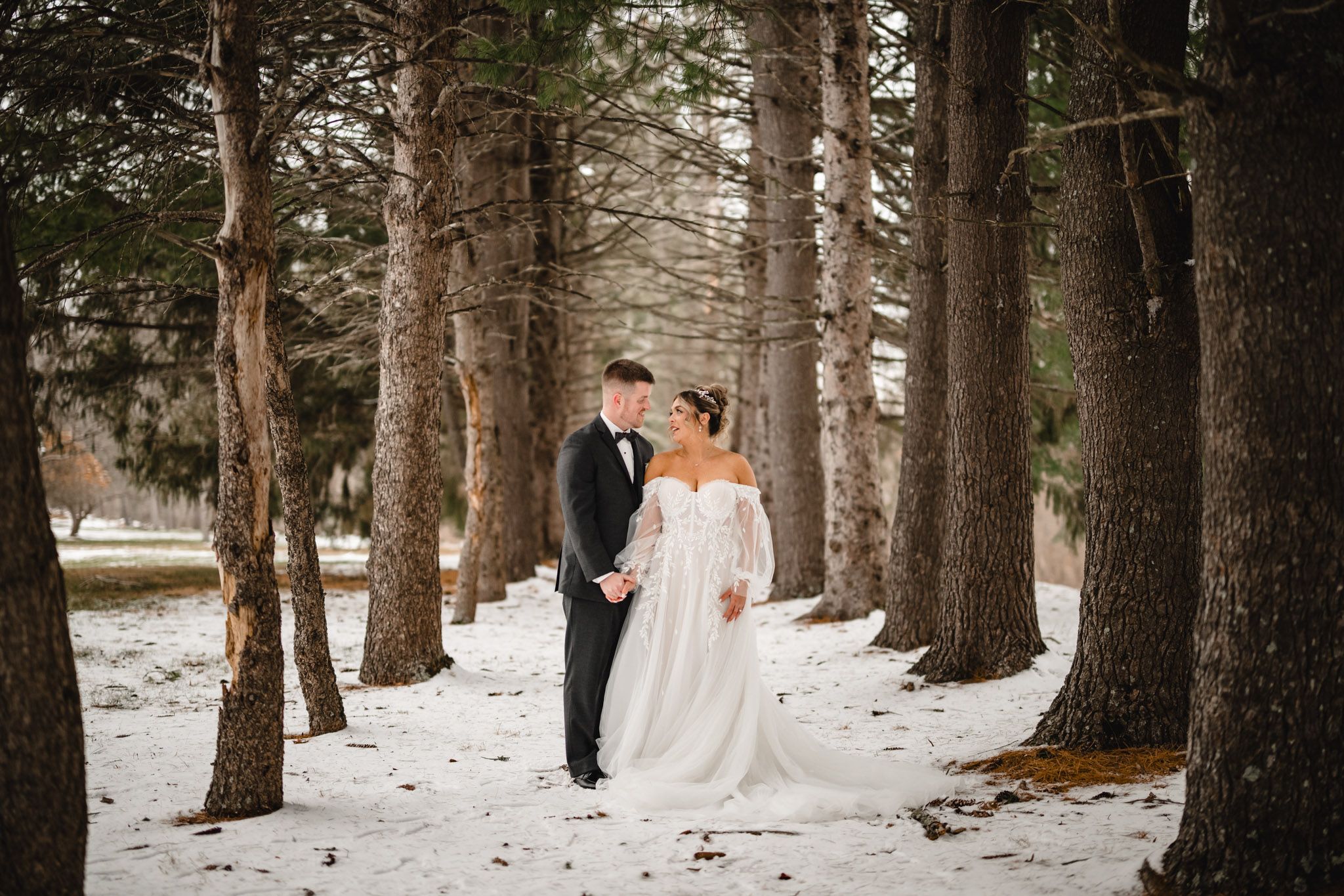 A romantic winter wedding photo captured in a snowy forest with tall pine trees creating a magical backdrop.