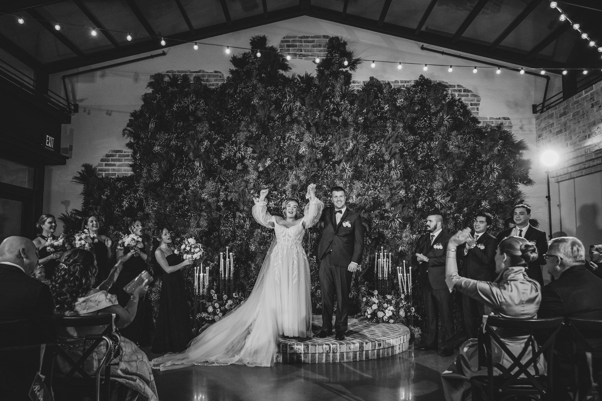A black and white photo of a wedding celebration with a large botanical wall backdrop.