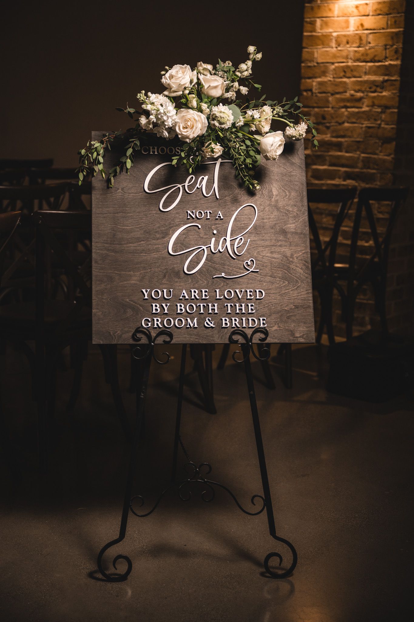 A wooden wedding sign decorated with white roses and greenery against exposed brick.