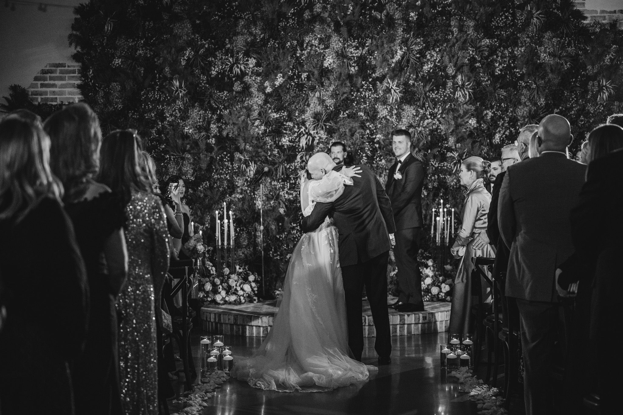 A black and white wedding ceremony photo showing a couple exchanging vows surrounded by guests.