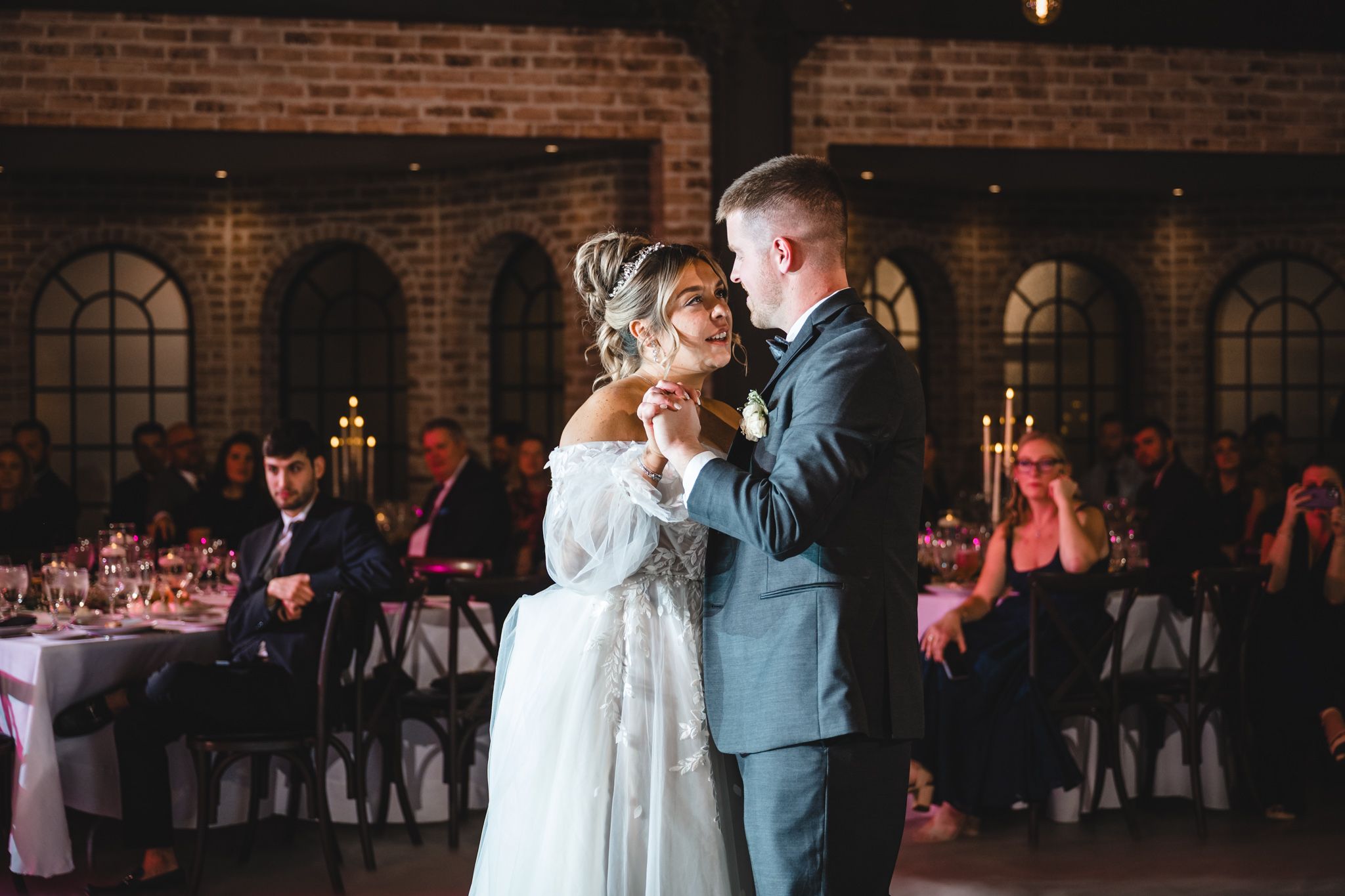 A romantic dance moment in a rustic venue with exposed brick walls and arched windows.