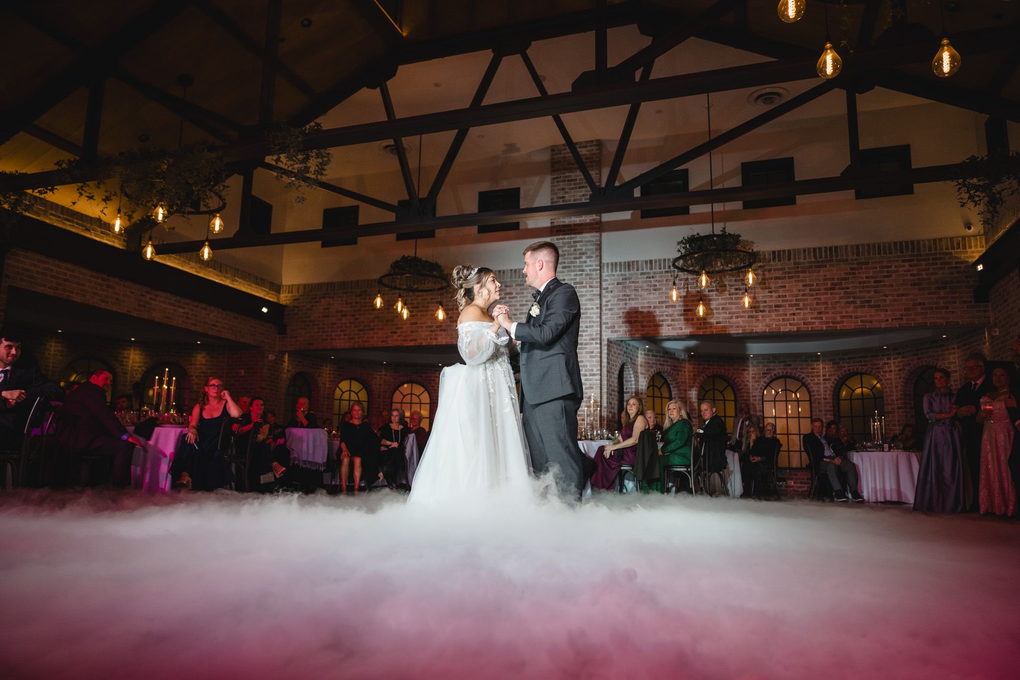 A couple shares their first dance on a misty dance floor in an elegant brick-walled reception venue with hanging lights.