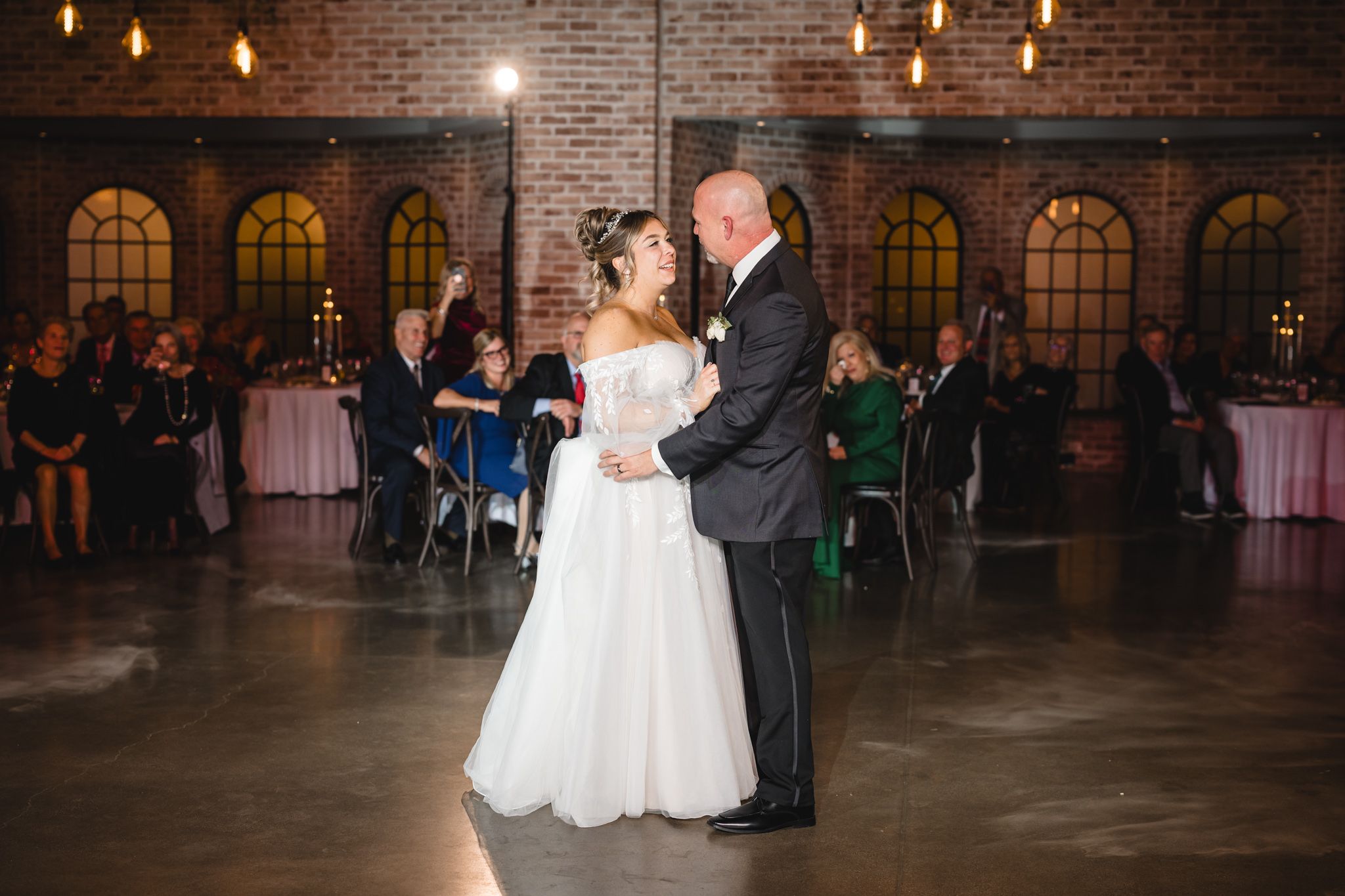 A wedding couple dances in an elegant venue with brick walls and arched windows.