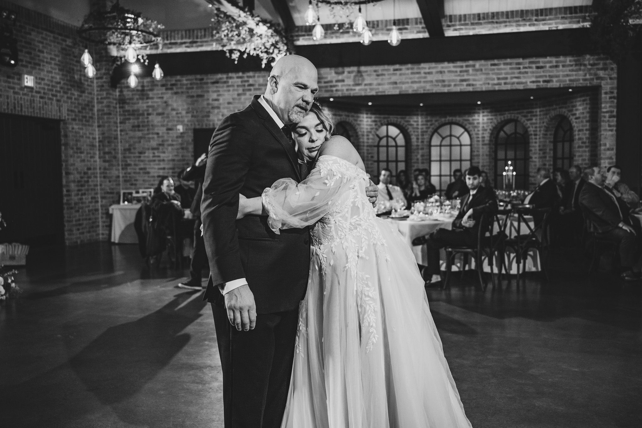 A black and white photo of a dance moment at a wedding reception with brick walls and arched windows.