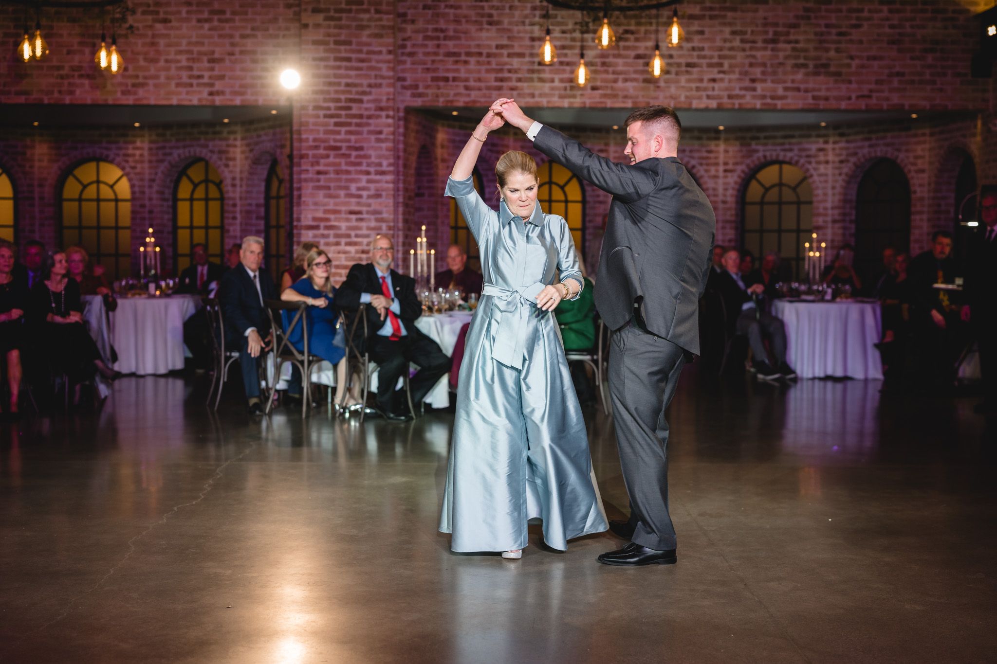A couple performs a dance twirl on a reception venue dance floor with pink-lit brick walls.
