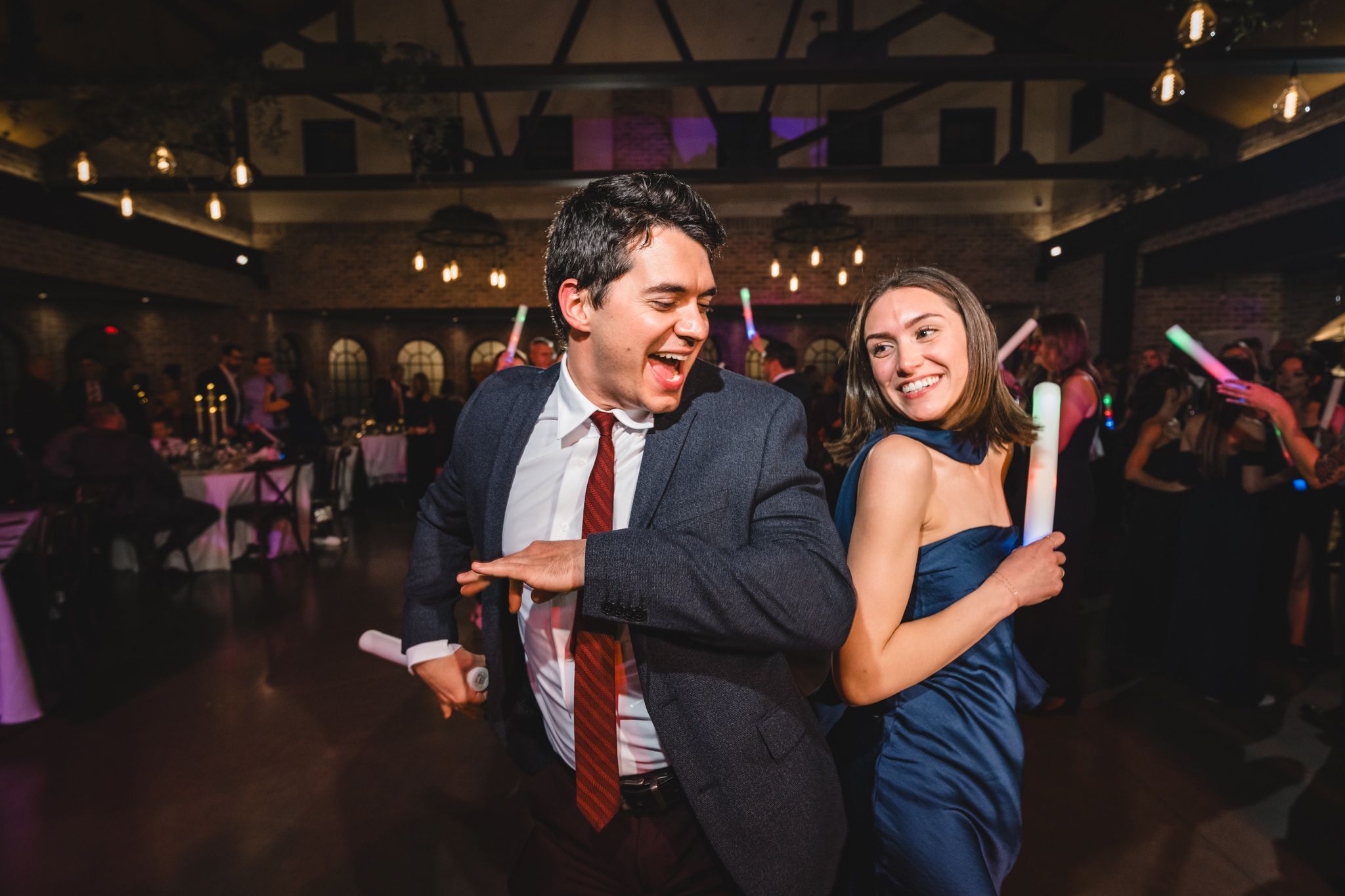 Wedding guests dancing energetically at a reception with exposed brick walls and mood lighting.
