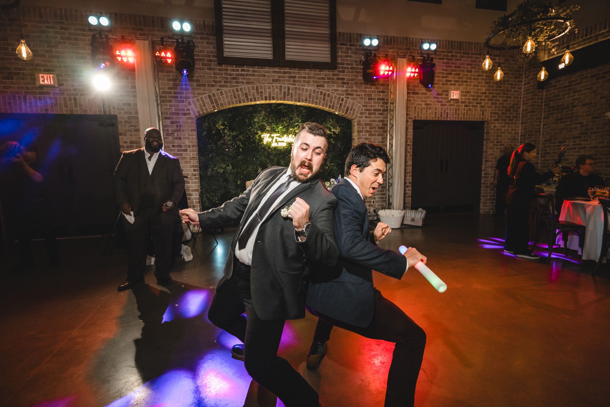 Two guests perform an energetic dance move under purple lighting at a wedding reception.