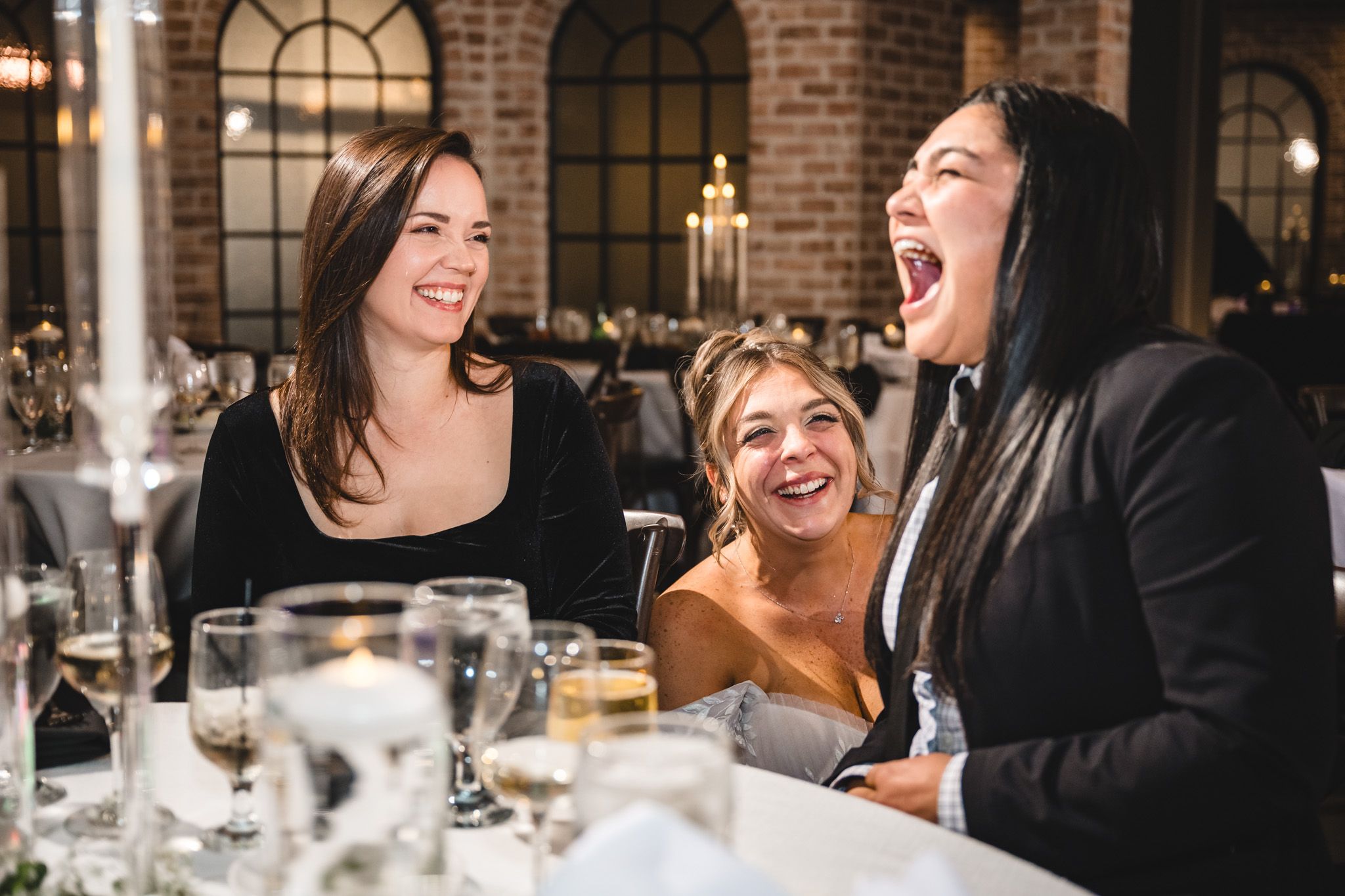Three wedding guests share a joyful moment of laughter at a reception table.