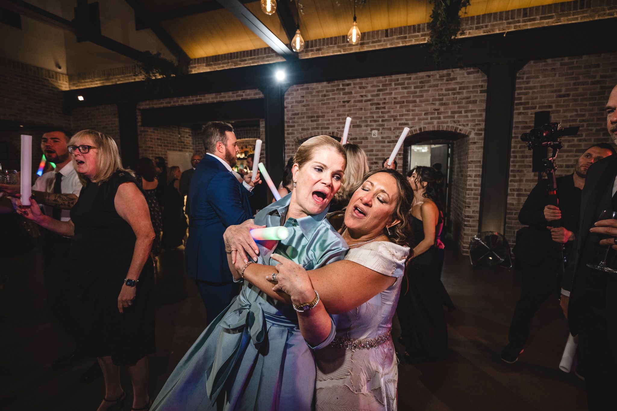 Wedding guests share a joyful dancing moment in a rustic brick venue with warm lighting.