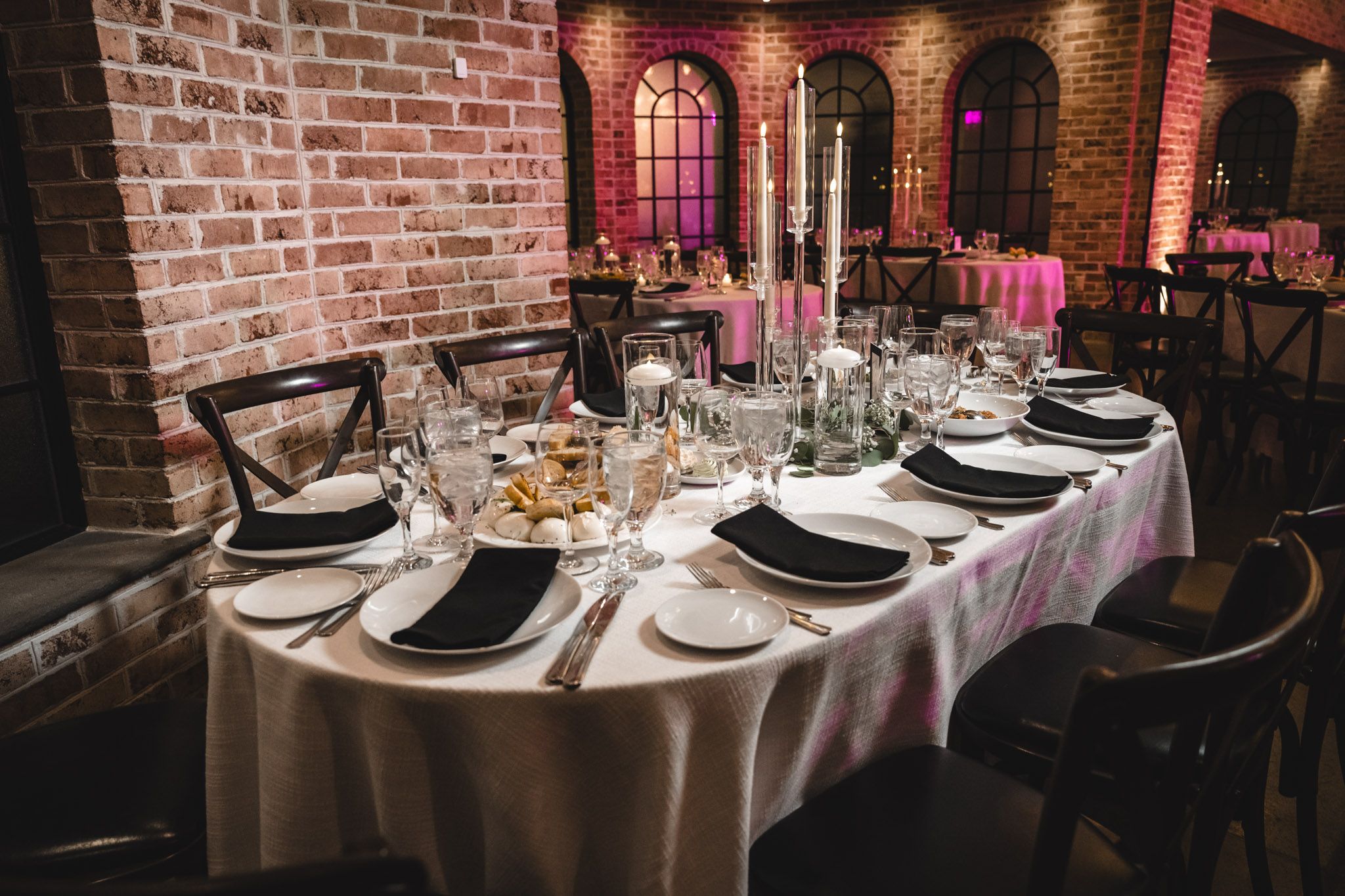 An elegant table setting with black chargers and tall candles in a brick-walled dining room.