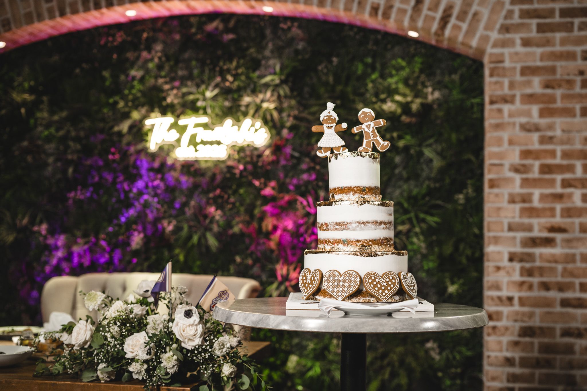 Wedding cake and dessert table display with floral decorations against a brick wall backdrop.