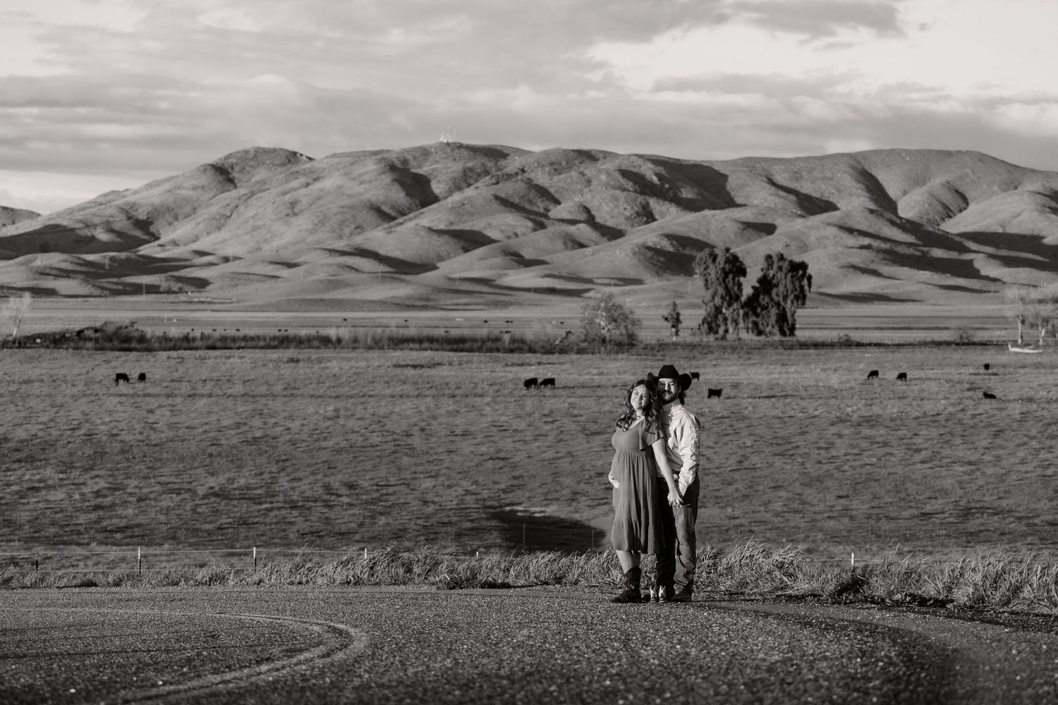 A couple embraces in a black and white landscape with rolling hills and mountains in the background.