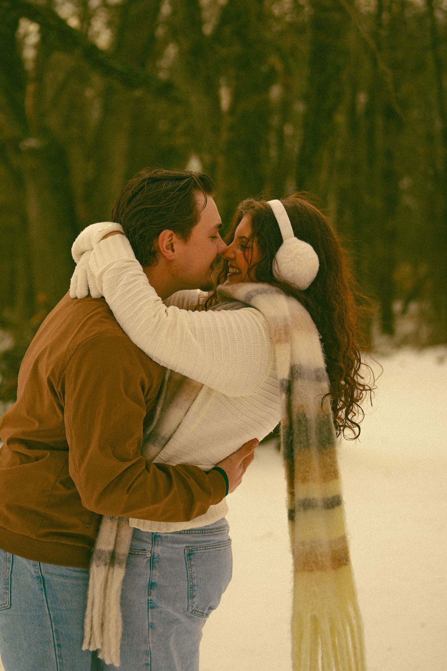 A couple in winter attire sharing an intimate moment in a snowy forest setting.