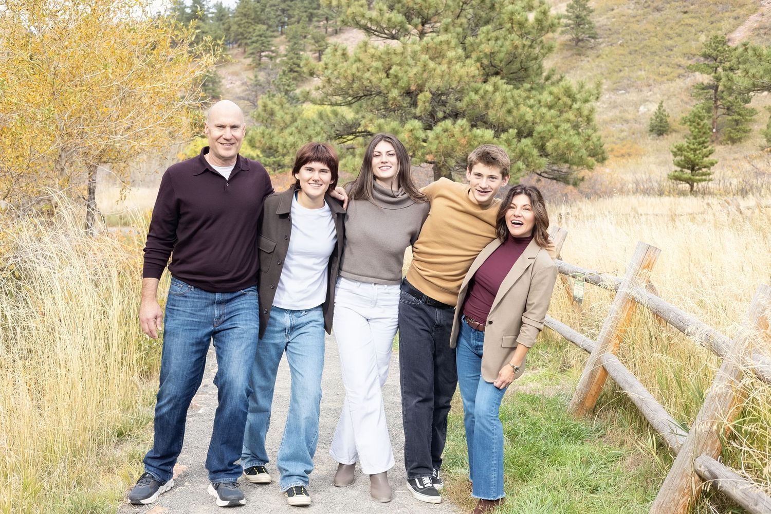 A group of people stand together outdoors in a natural setting with autumn grass and wooden fence in the background.