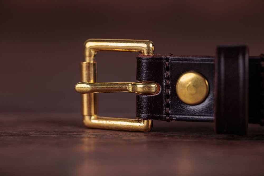 Black leather belt with gold brass buckle and rivet on wooden surface.