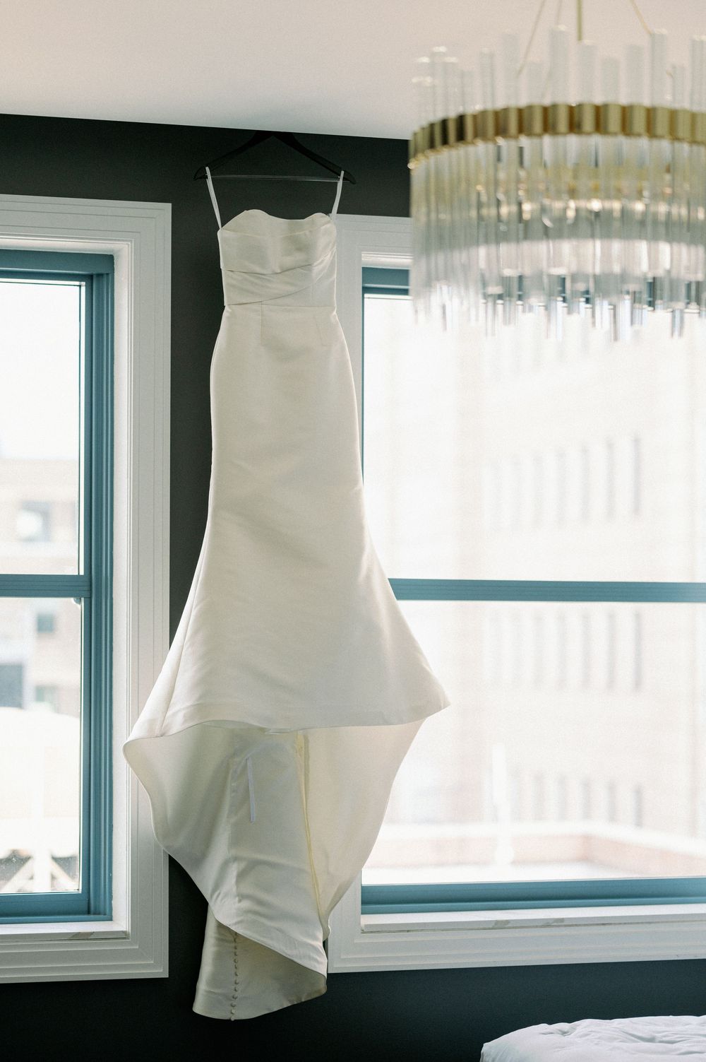 An elegant white mermaid wedding dress hangs by a window under a crystal chandelier in a sunlit room. - Fairlie Chicago Wedding