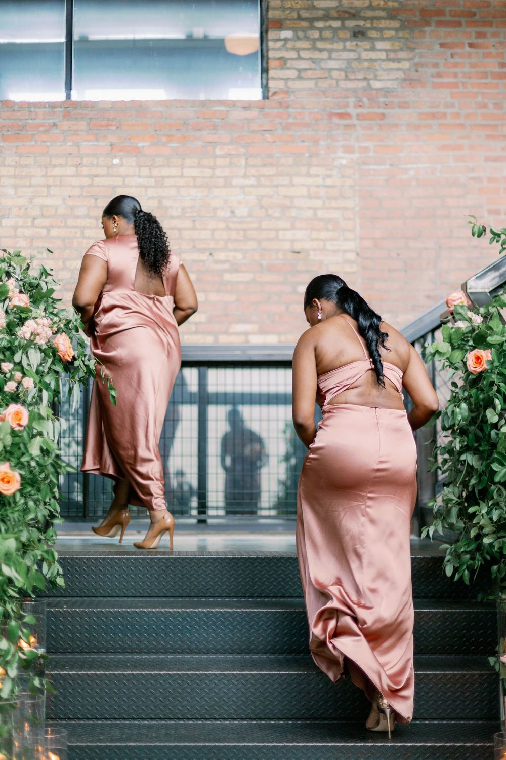 Two figures in blush pink satin dresses ascend floral-decorated stairs against a brick wall. - Fairlie Chicago Wedding