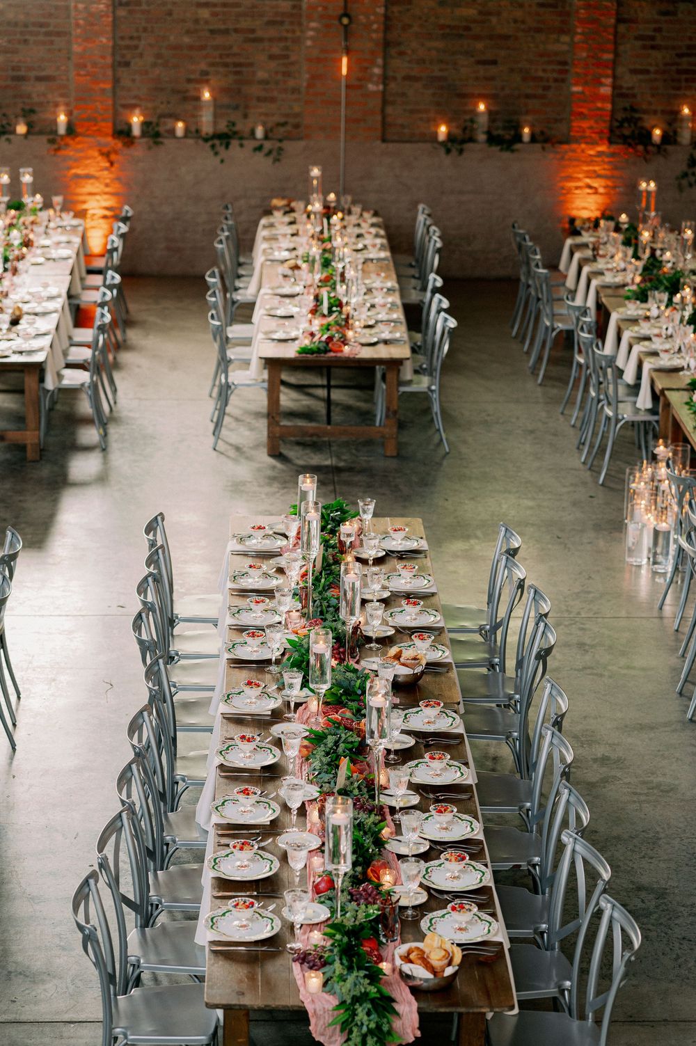 Long rustic dining tables with greenery garlands and place settings in an industrial brick venue with warm lighting. - Fairlie Chicago