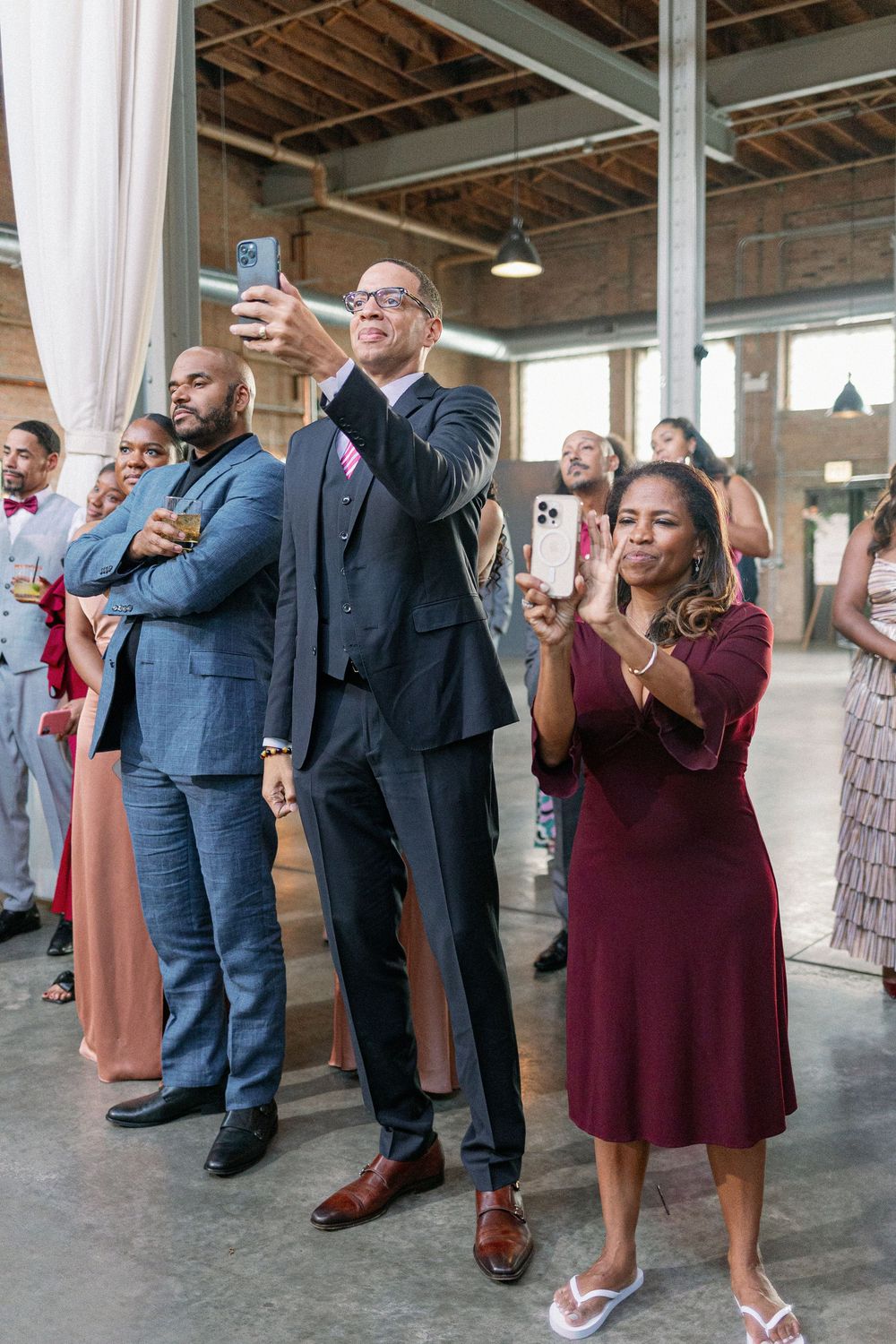 Wedding guests in formal attire gather in a rustic brick venue space for group photos. - Fairlie Chicago Wedding