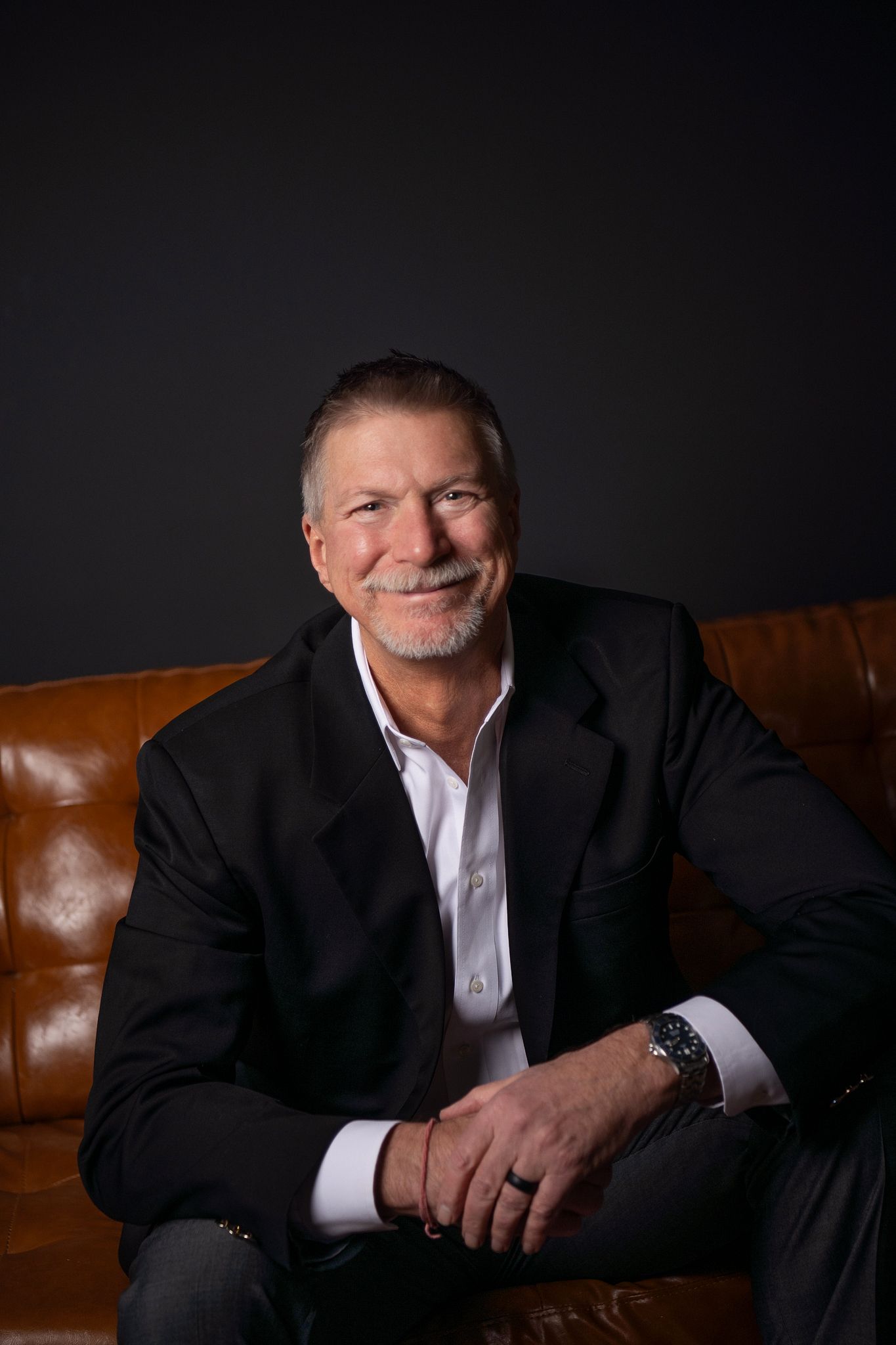 Professional portrait series of person in dark suit against black background seated on brown leather couch.