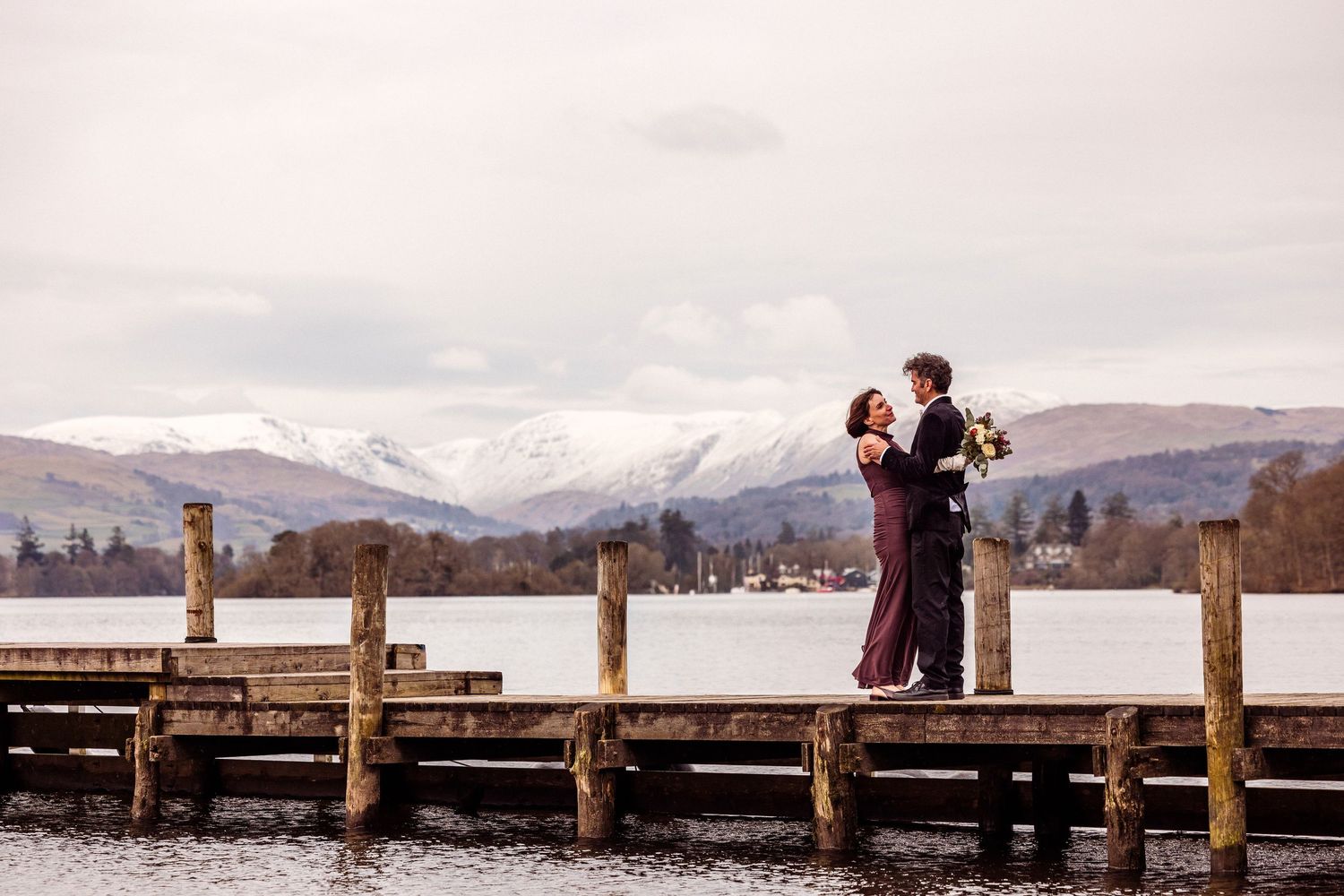 A couple stands together on a jetty at Storrs Hall with snow covered mountains in the background