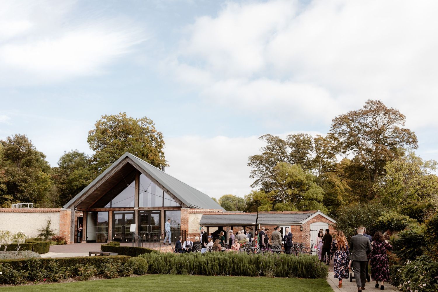 Panoramic view of a rustic barn wedding venue with guests gathering outside during a sunny afternoon celebration.