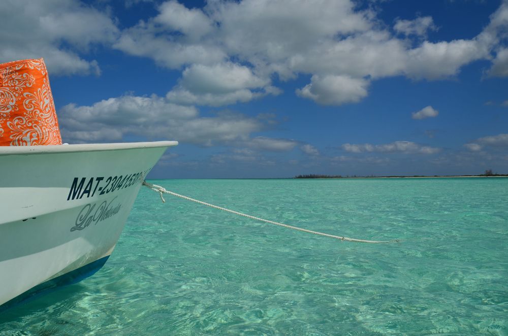A small white fishing boat floats in crystal‑clear turquoise water under a bright blue sky, anchored just offshore with a soft breeze